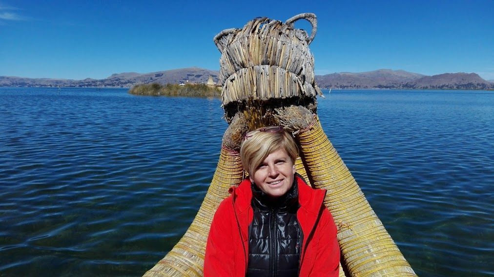 Mujer con abrigo rojo en una barca de totora en el lago Titicaca, agua azul y cielo, montañas al fondo.