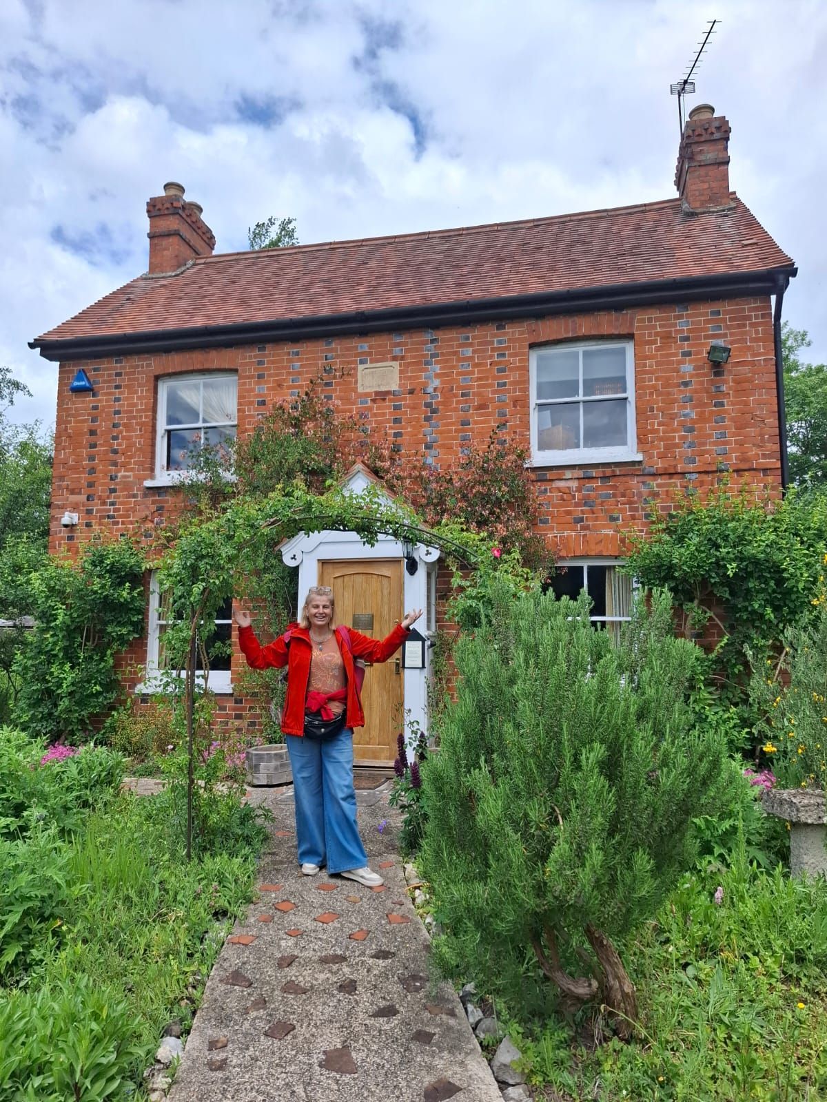 Persona con los brazos extendidos frente a una cabaña de ladrillo rojo con un jardín de flores.