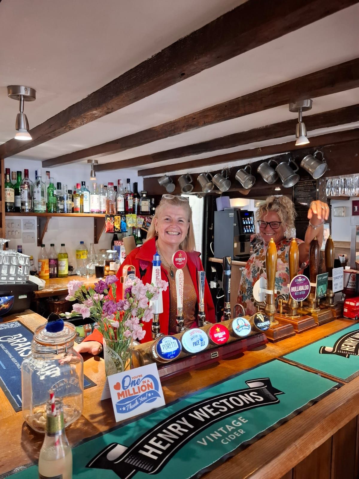 Dos personas detrás de la barra de un pub con grifos y botellas de cerveza, flores y carteles.
