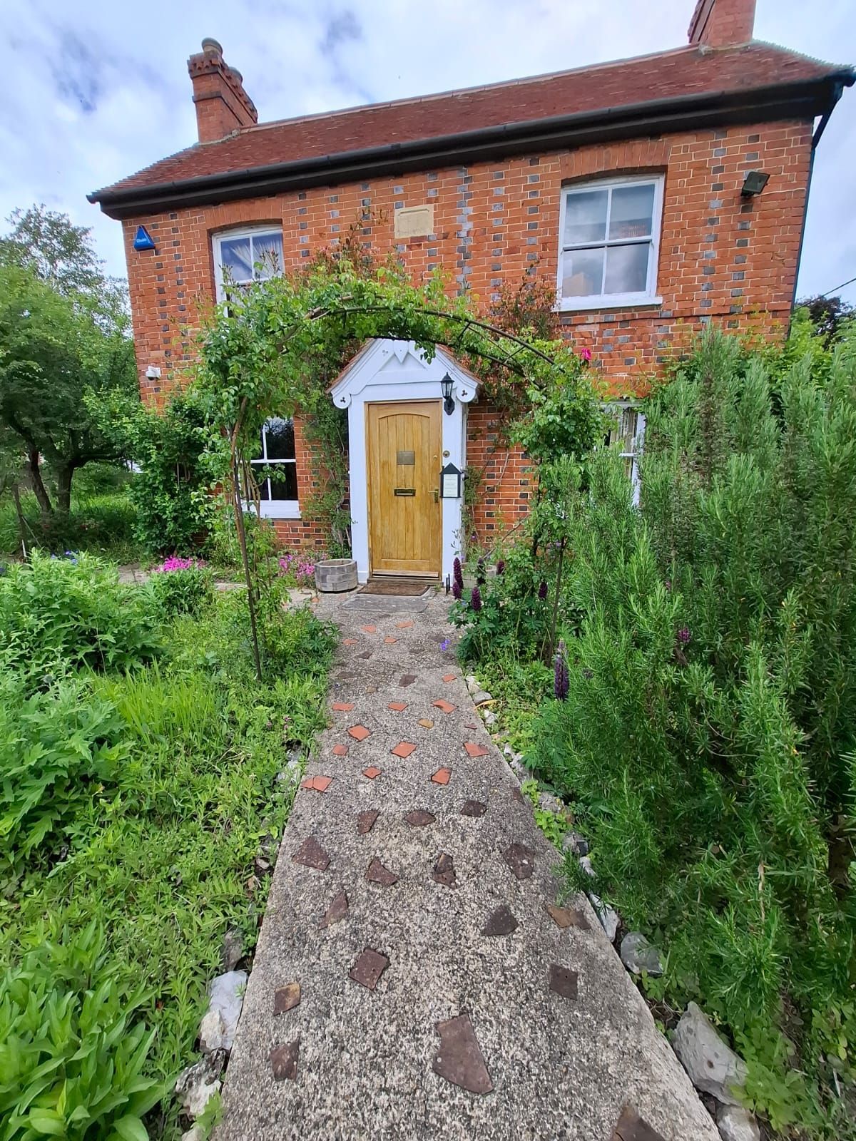 Cabaña de ladrillo con un camino de piedra que conduce a una puerta de madera, rodeada de exuberante vegetación y flores.