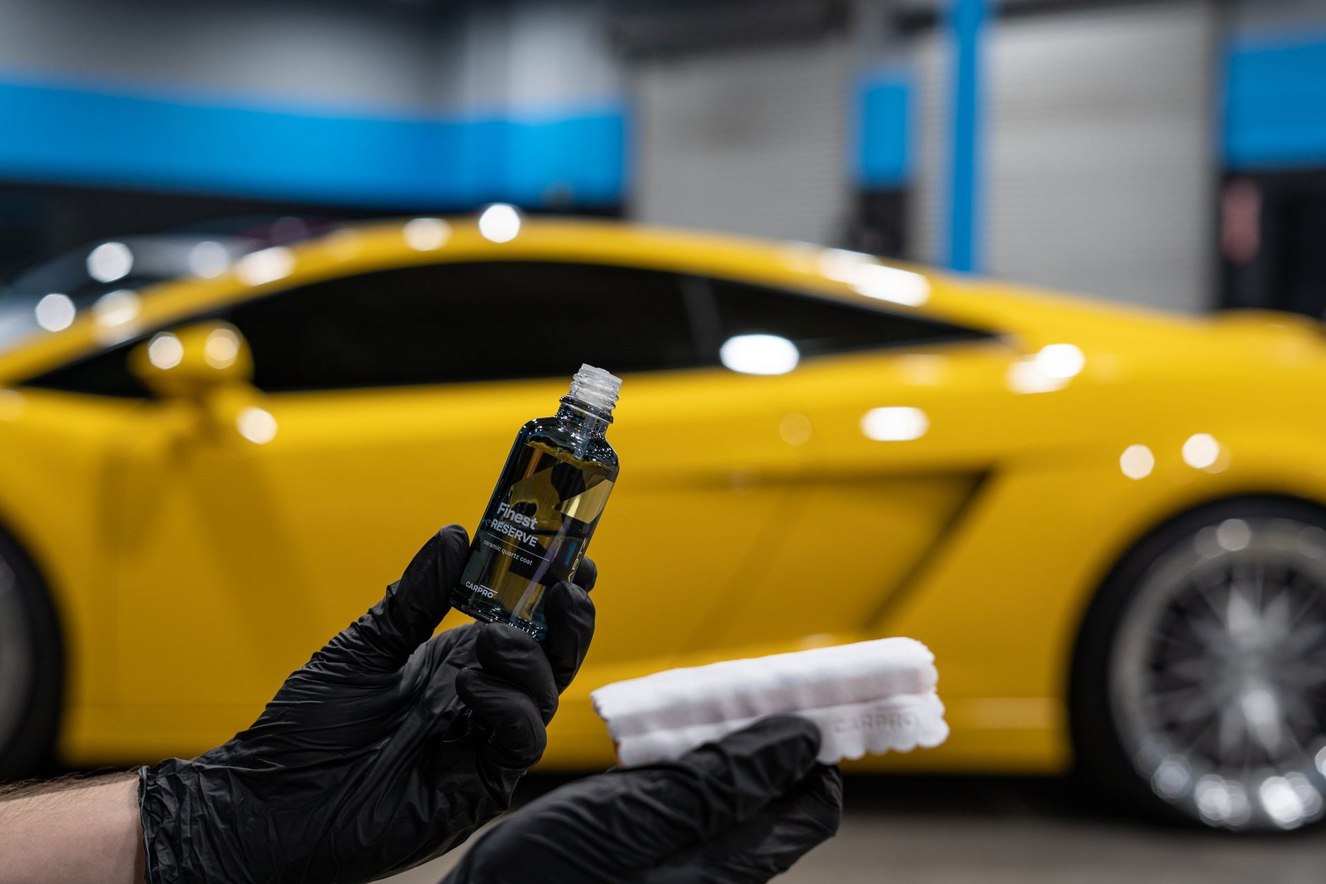 A person is holding a bottle of ceramic coating in front of a yellow car.