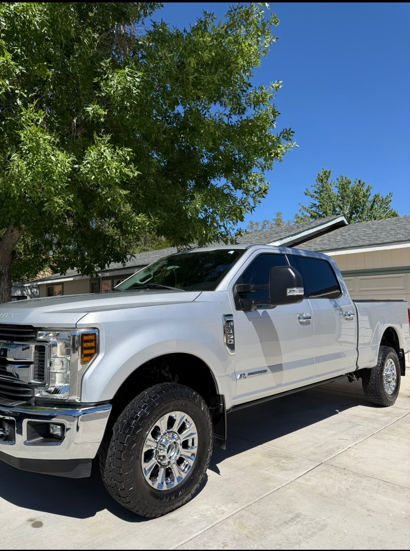 Silver Ford F-350 pickup truck parked on a paved driveway with a house and tree in the background.