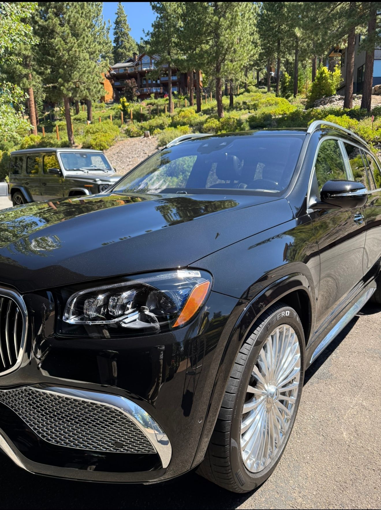 Black Mercedes GLS SUV parked on a driveway, shiny wheels. Mountains and trees in the background.