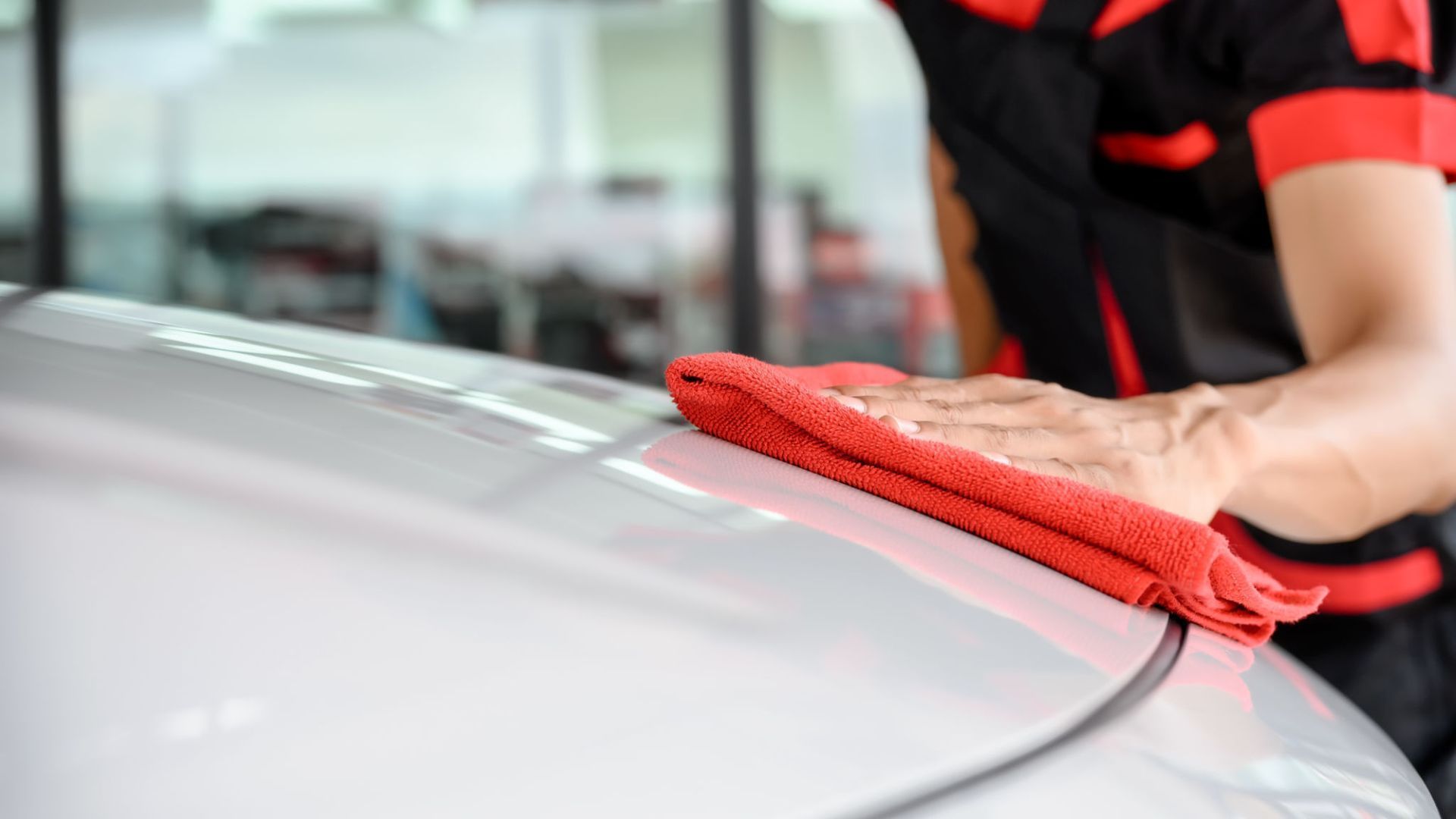 A man is cleaning the hood of a car with a red towel.