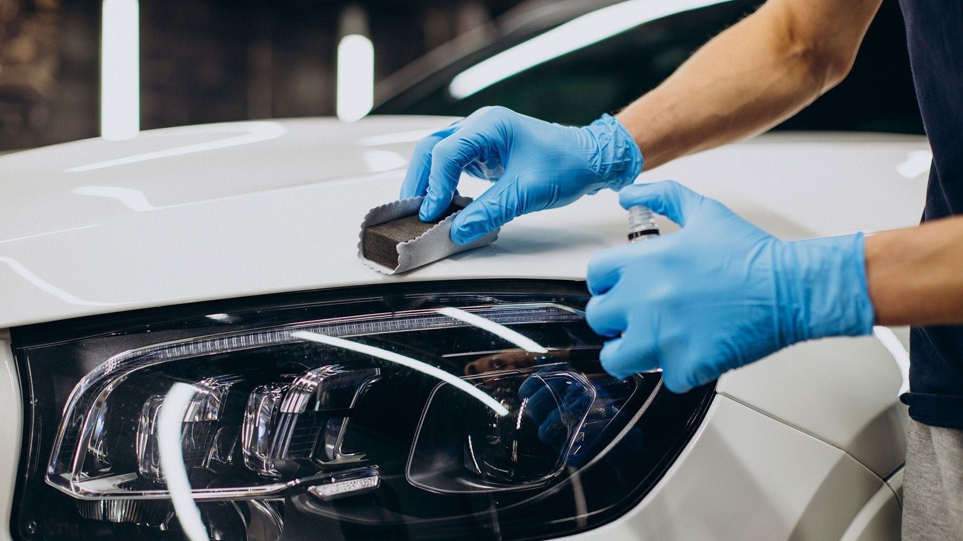 A person wearing blue gloves is polishing the headlights of a white car.