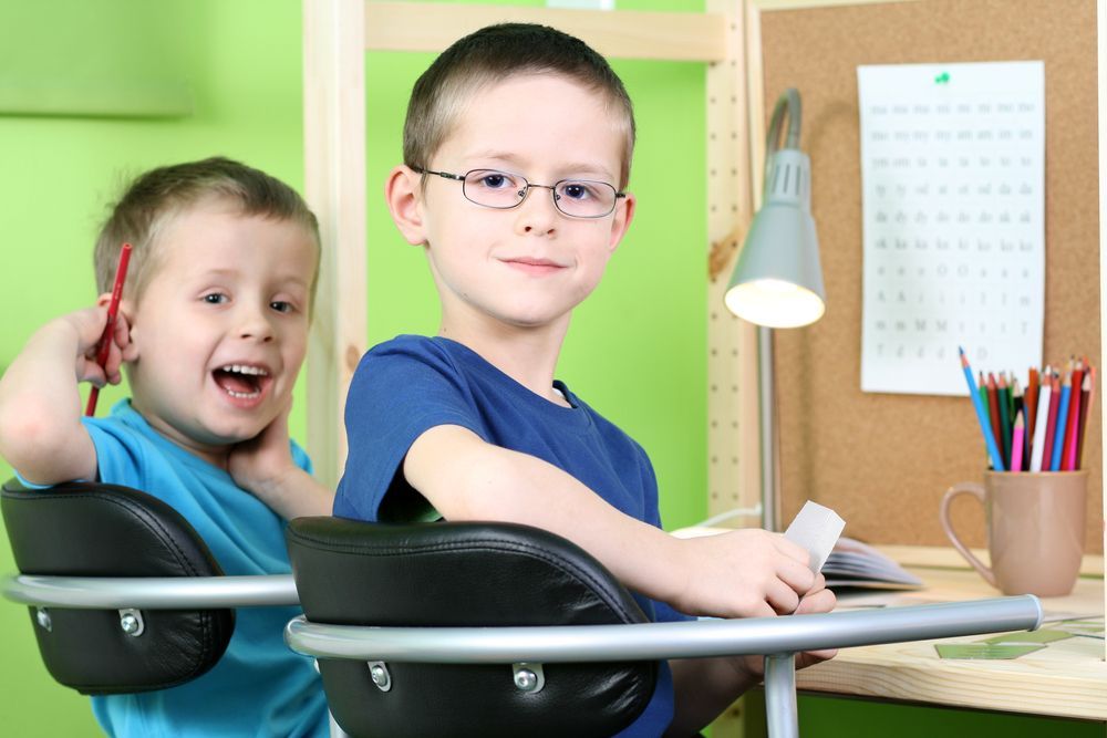 Two boys at a desk, one wearing glasses and smiling, other excited with pencil. Green wall, desk lamp.