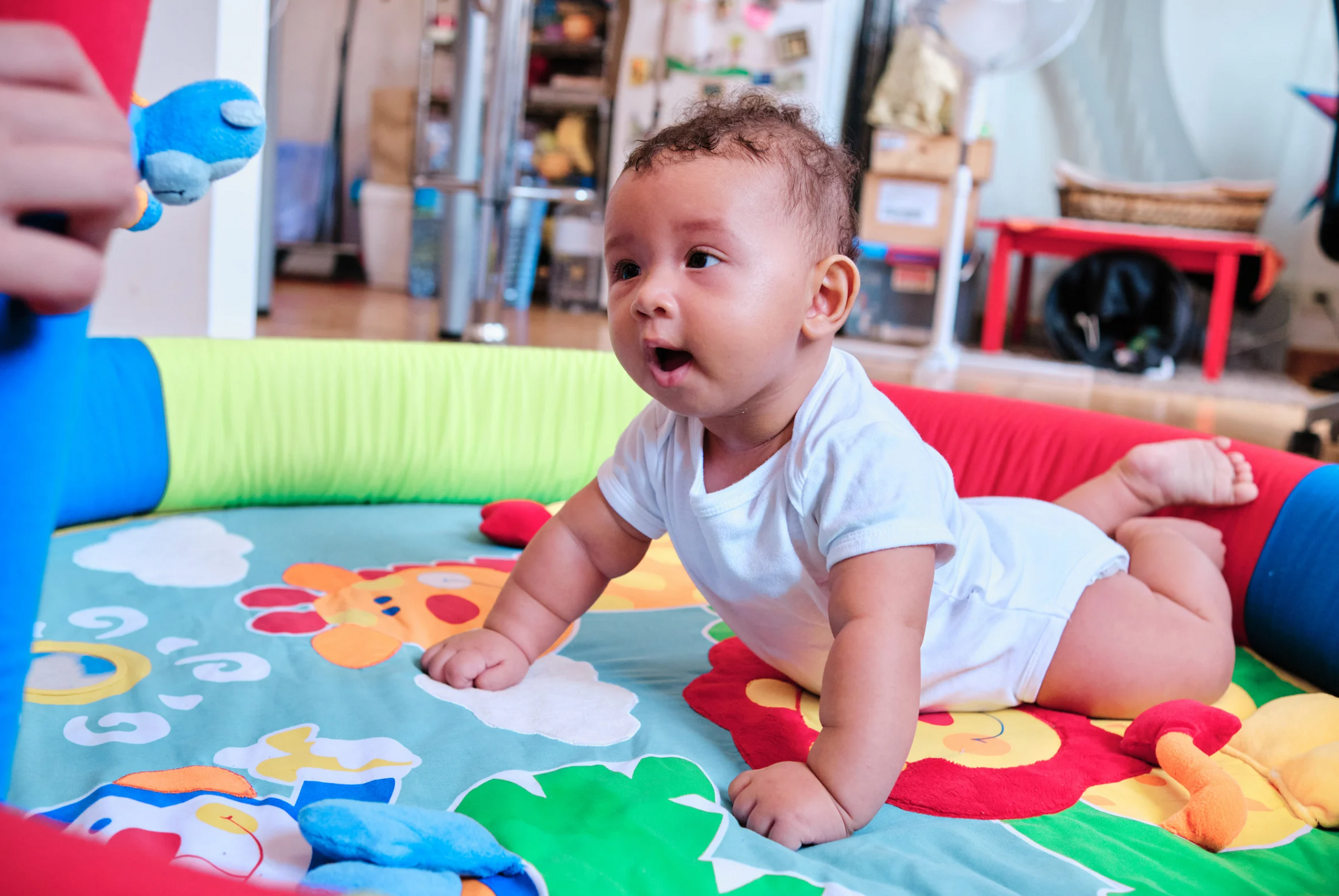 Baby on a colorful playmat, looking up with mouth open; a toy is held above him.