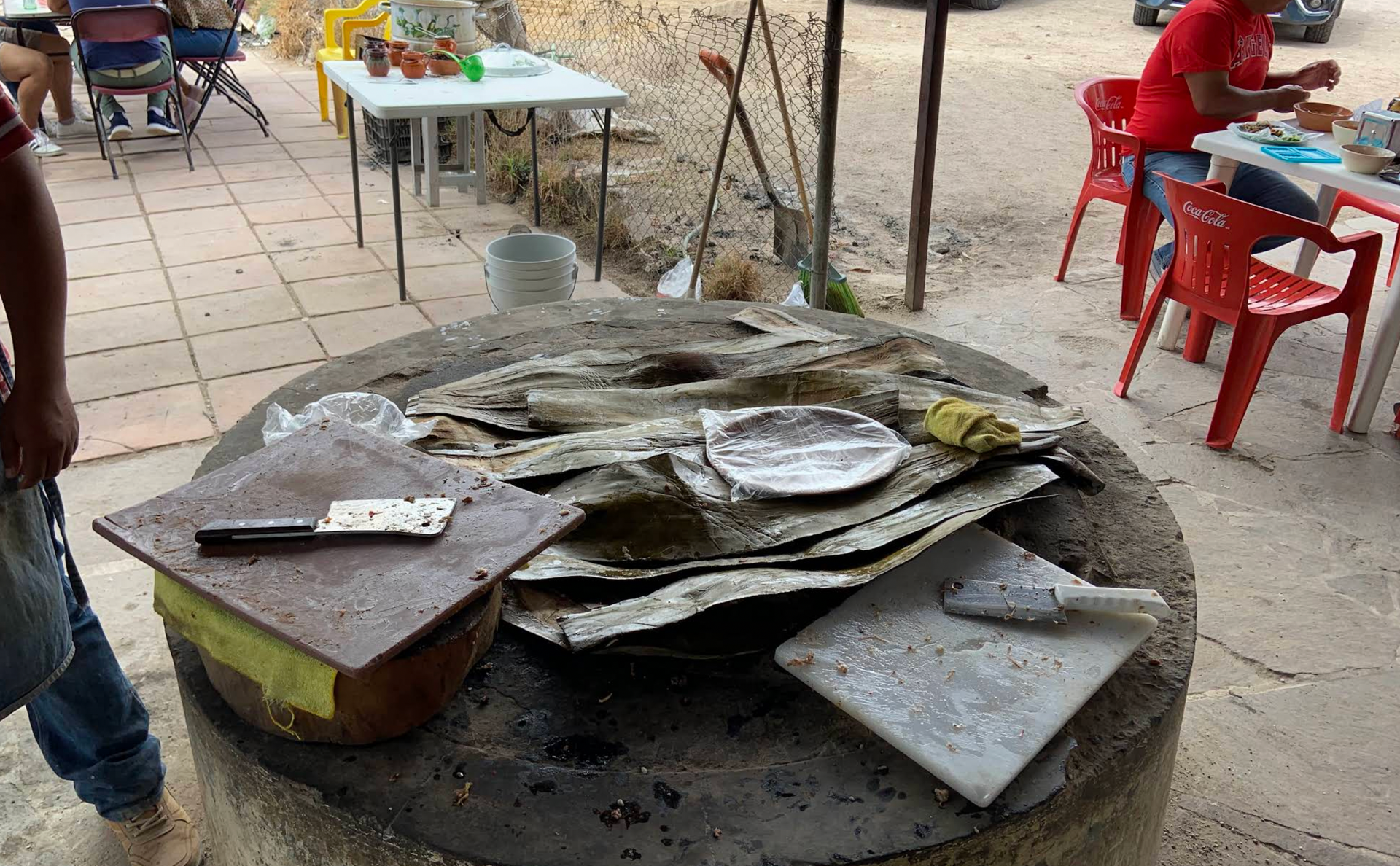 Gran parrilla al aire libre con tortillas y preparación de alimentos. Personas sentadas a las mesas al fondo.