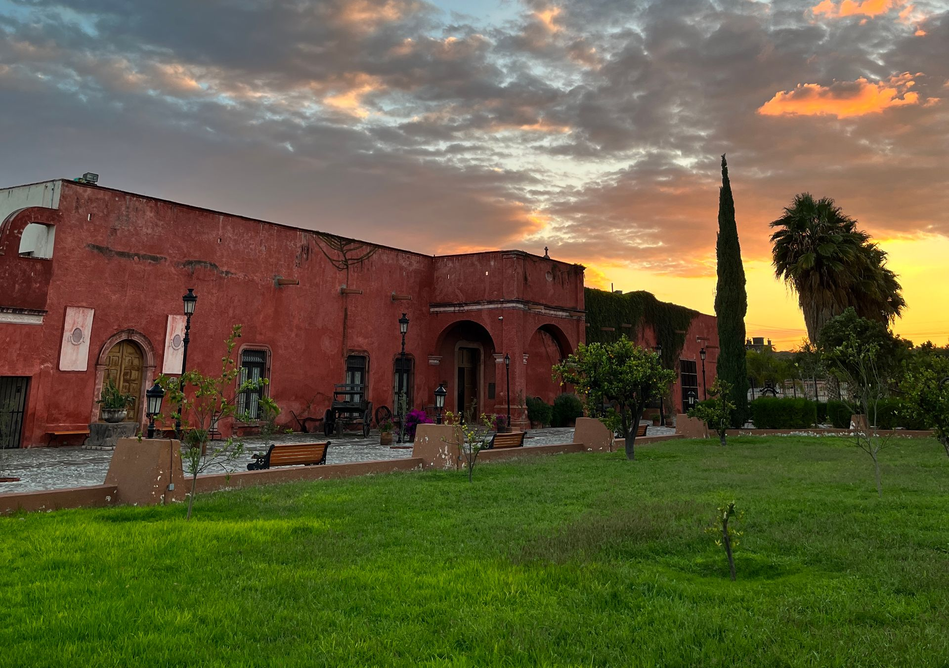 Edificio de ladrillo rojo con arcos, césped y árboles altos bajo un cielo de atardecer lleno de color.
