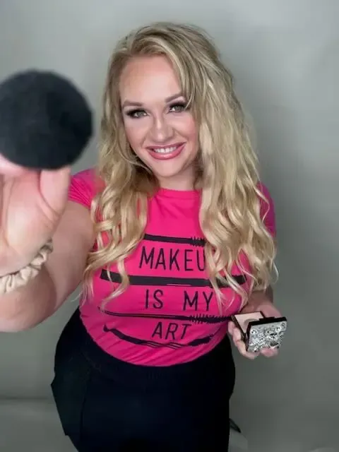 A woman wearing a pink t-shirt that says `` makeup is my art '' is holding a makeup brush.