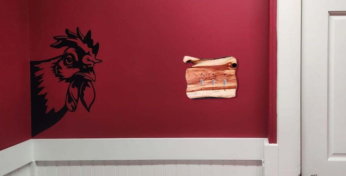 A red wall with a rooster and a rustic wood 3 toggle light switch in juniper.