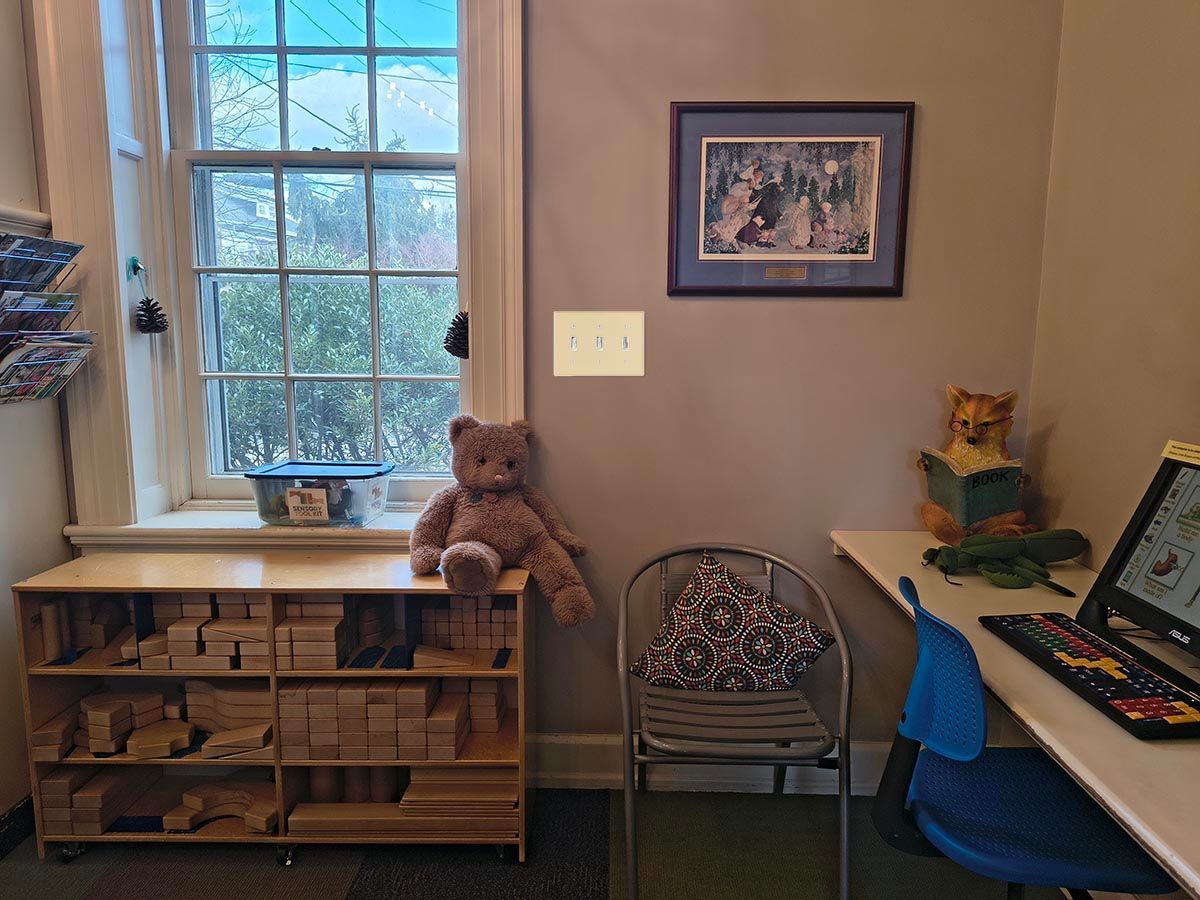 Room with window, wooden shelves, a bear, chair, desk with laptop, and framed picture.