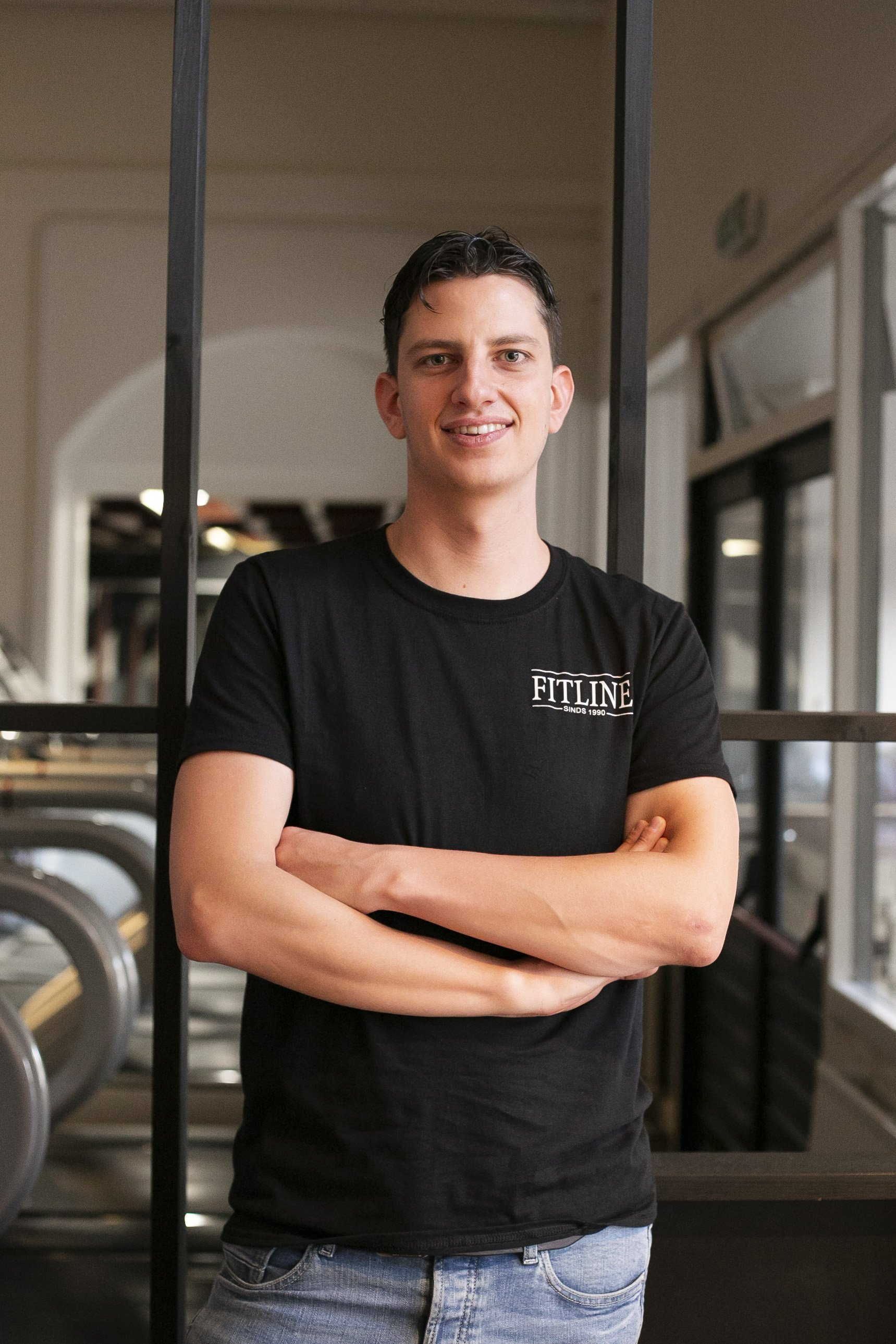 Een man in een zwart shirt staat met zijn armen over elkaar in een sportschool.