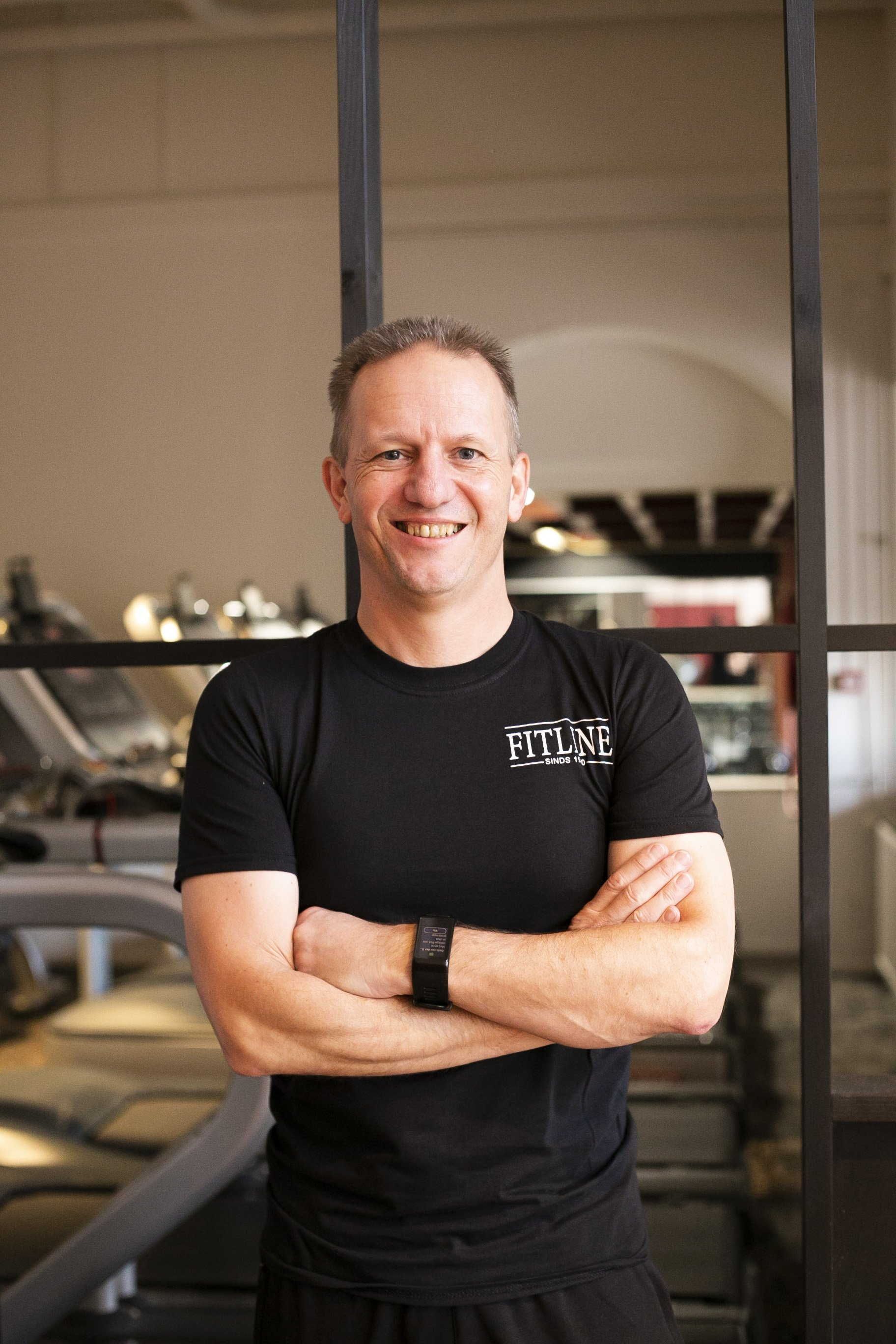 Een man in een zwart shirt staat in een sportschool met zijn armen over elkaar.