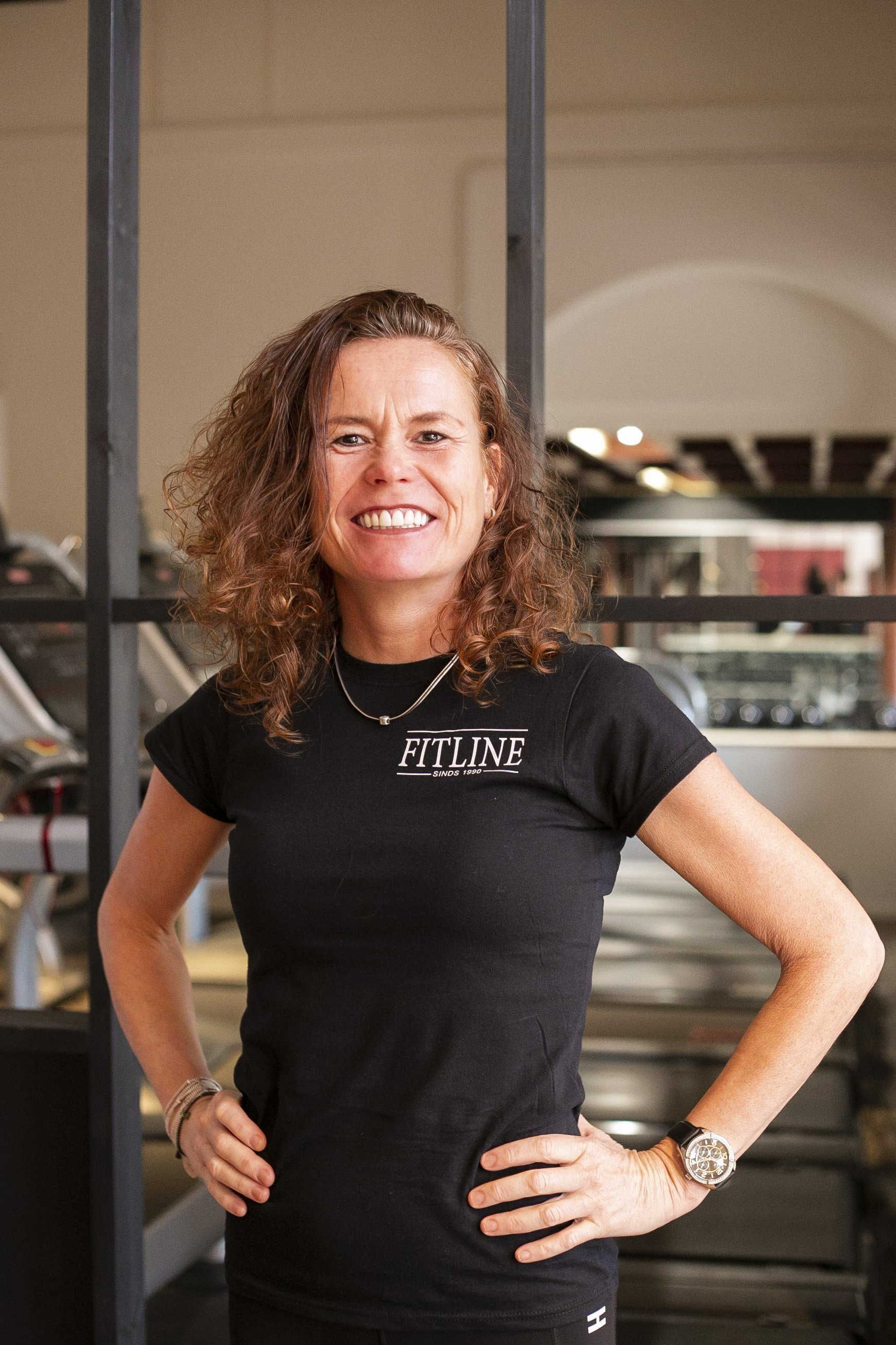 Een vrouw in een zwart shirt staat in een sportschool met haar handen in haar zij.
