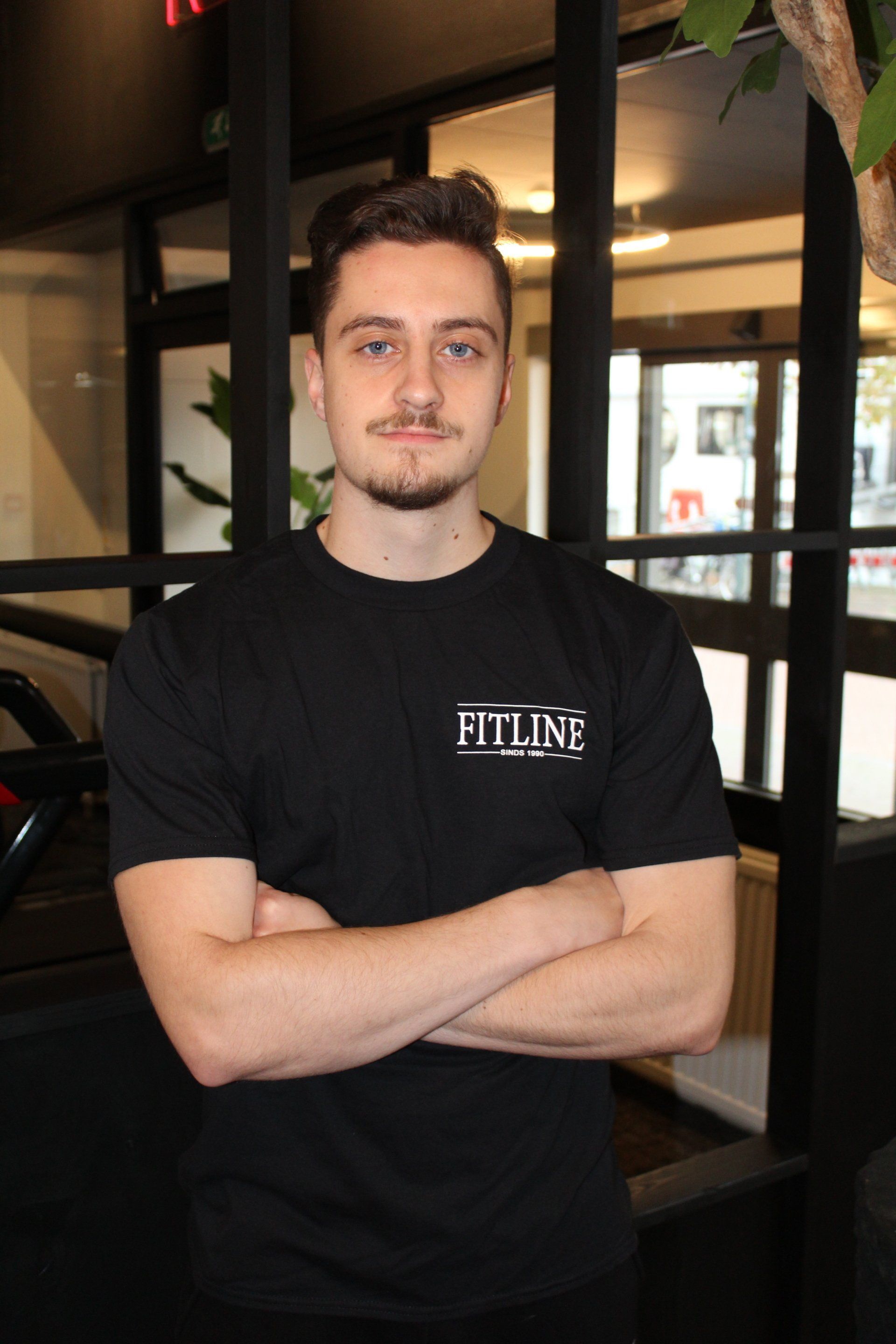 Een man in een zwart t-shirt staat met zijn armen over elkaar in een sportschool.