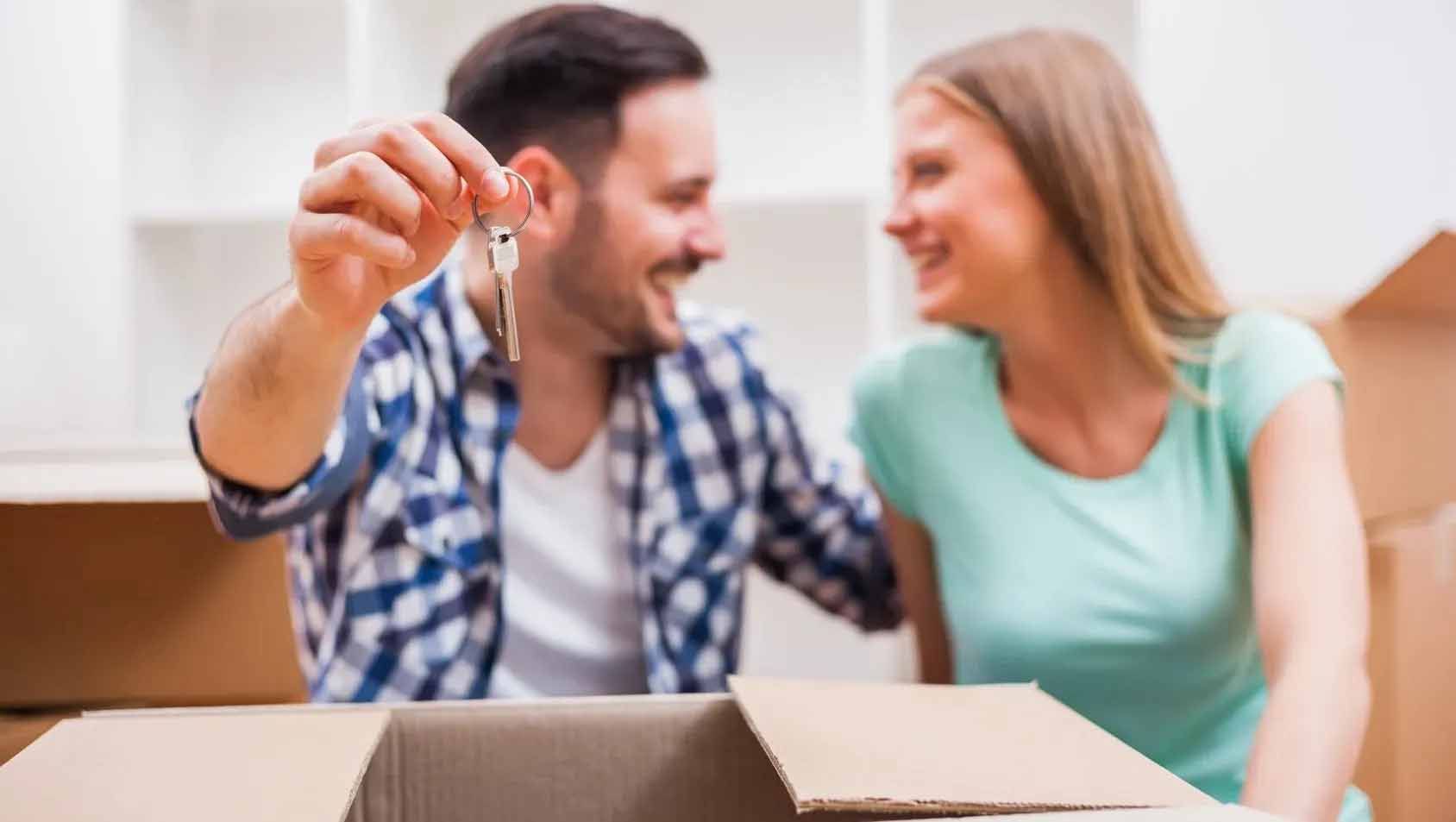 A man and a woman are sitting in a cardboard box holding keys to their new home.