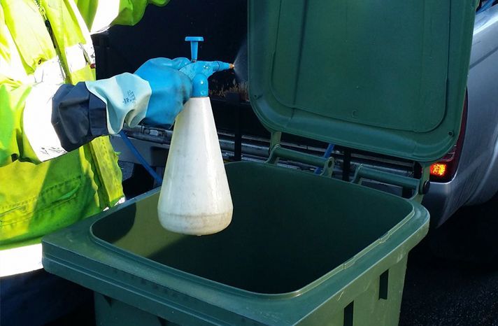 A man is spraying a green trash can with a spray bottle.