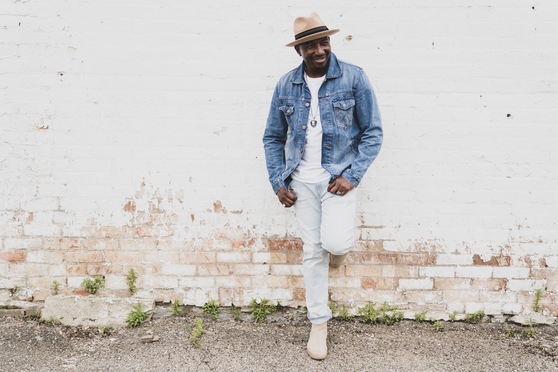 Corey Tucker in a denim jacket and hat is standing in front of a white brick wall.