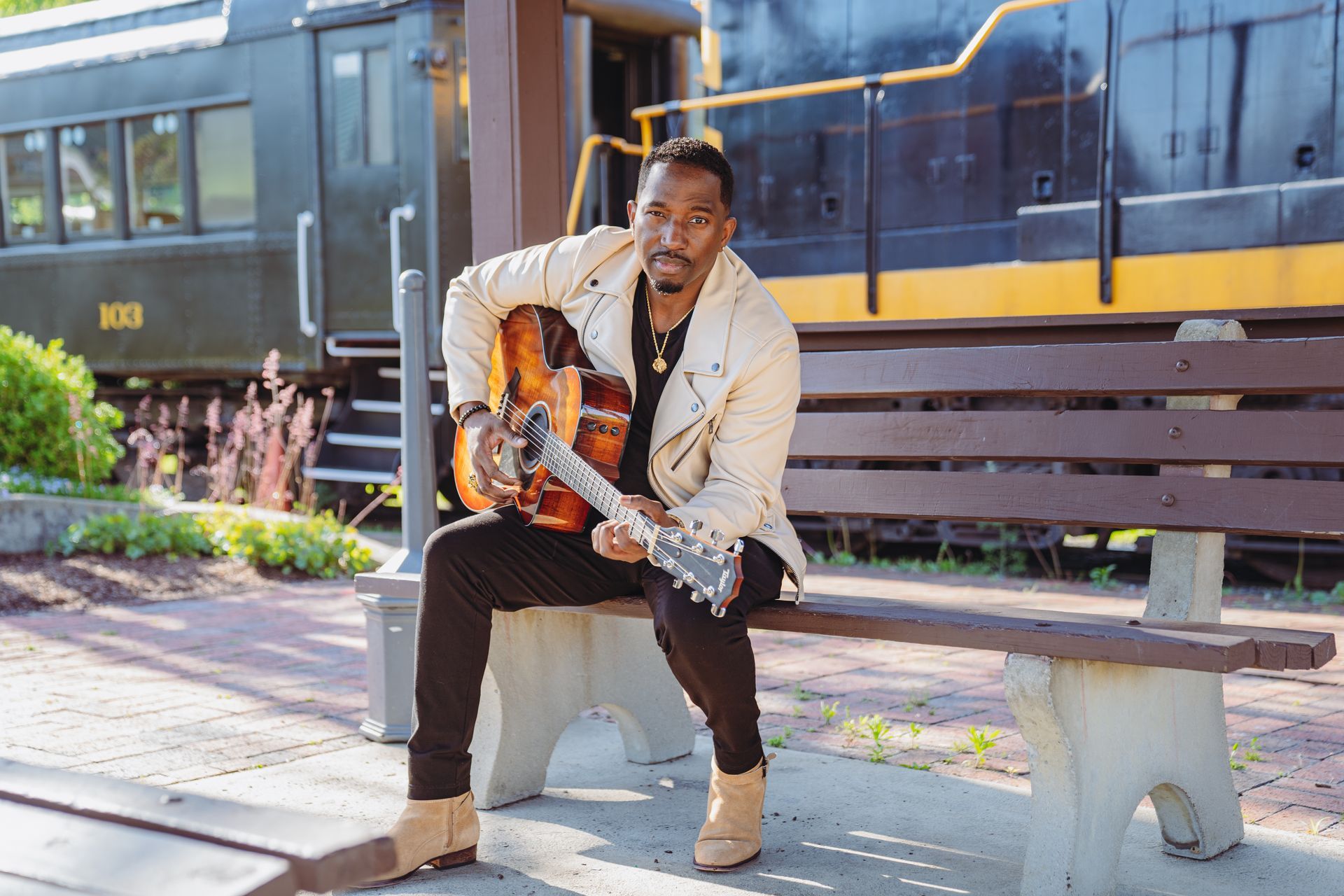 Corey Tucker is sitting on a bench holding a guitar in front of a train.