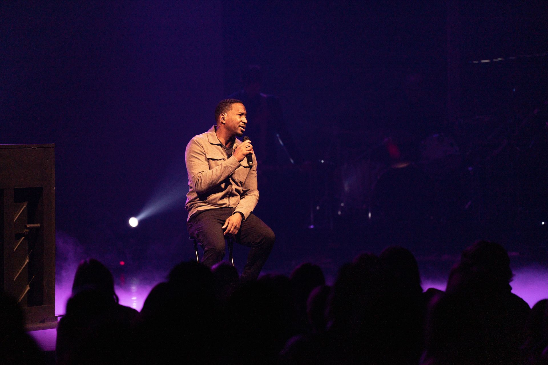 Corey Tucker is sitting on a stage holding a microphone in front of a crowd.
