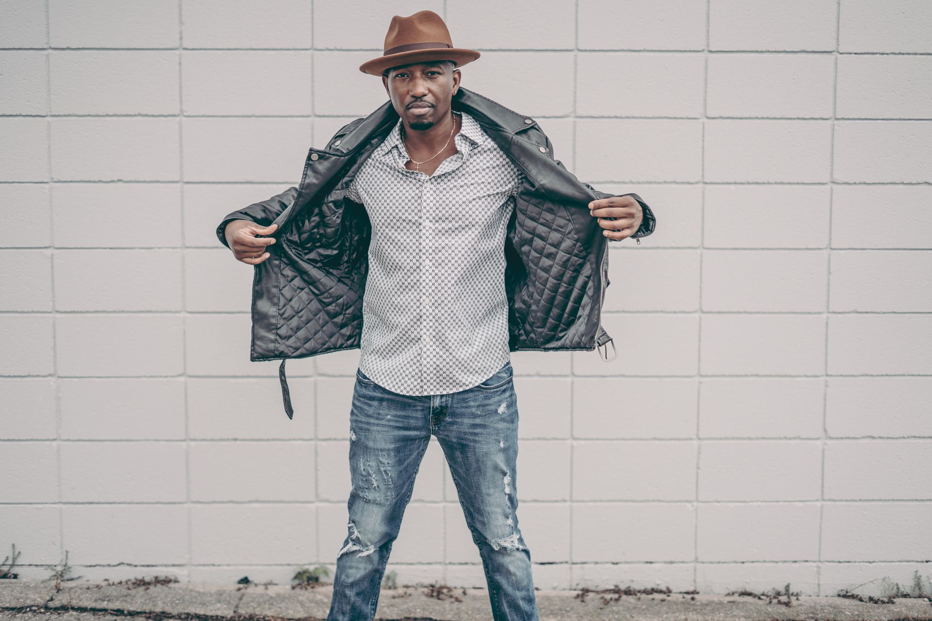 Corey Tucker wearing a hat and a leather jacket is standing in front of a white brick wall.