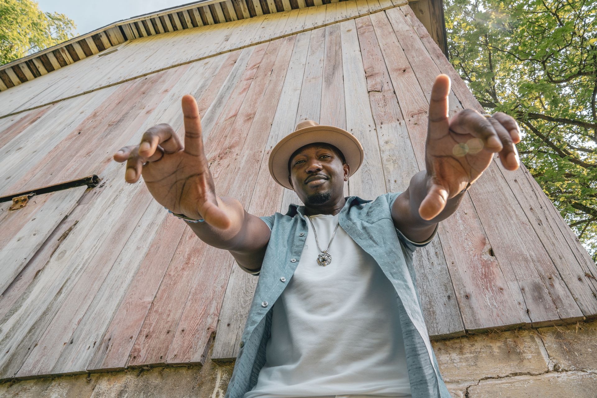 Corey Tucker in a hat is standing in front of a wooden building with his arms outstretched.
