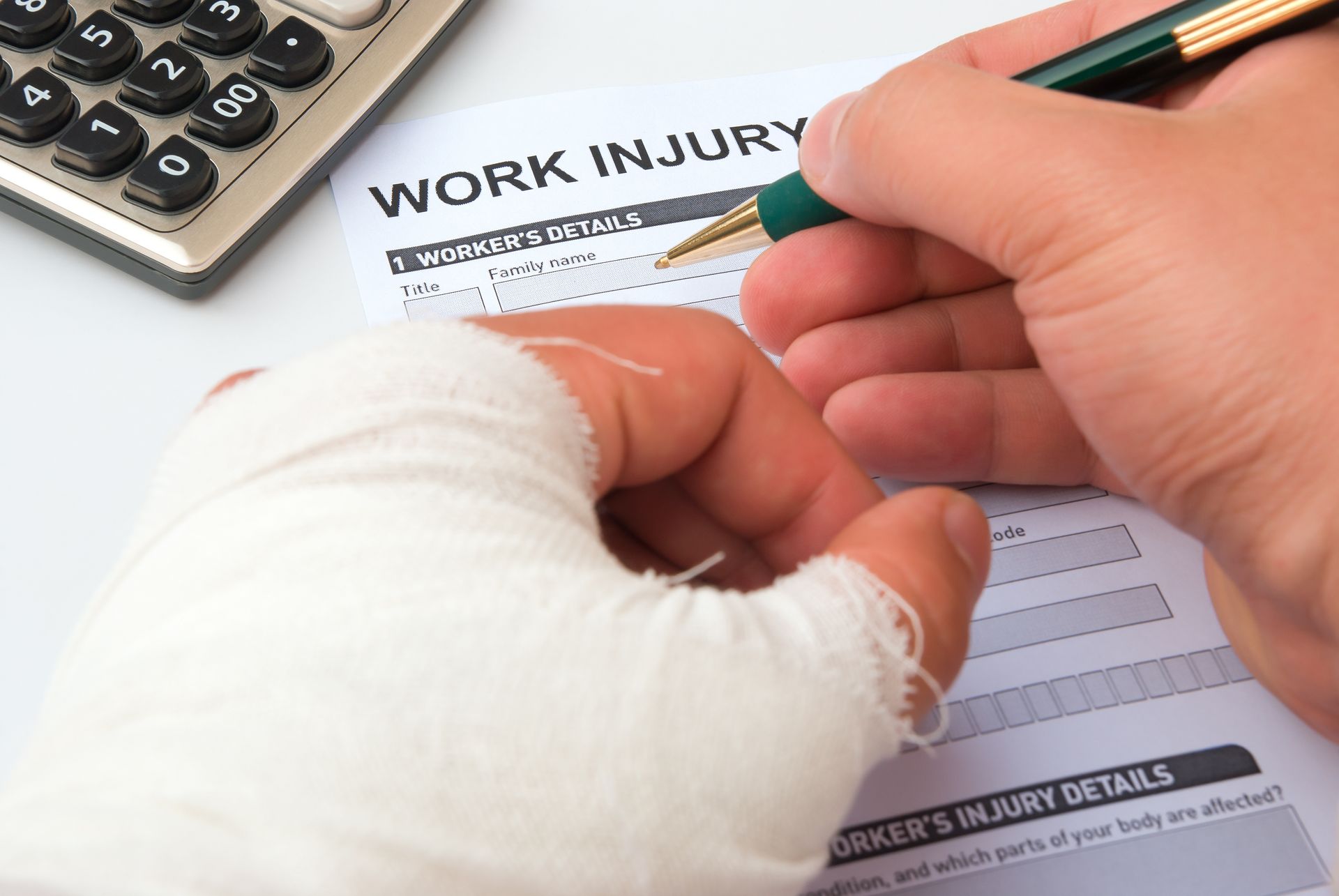 Close-up of bandaged hand filling out “Work Injury” form with pen next to calculator. Close-up of bandaged hand filling out “Work Injury” form with pen next to calculator.