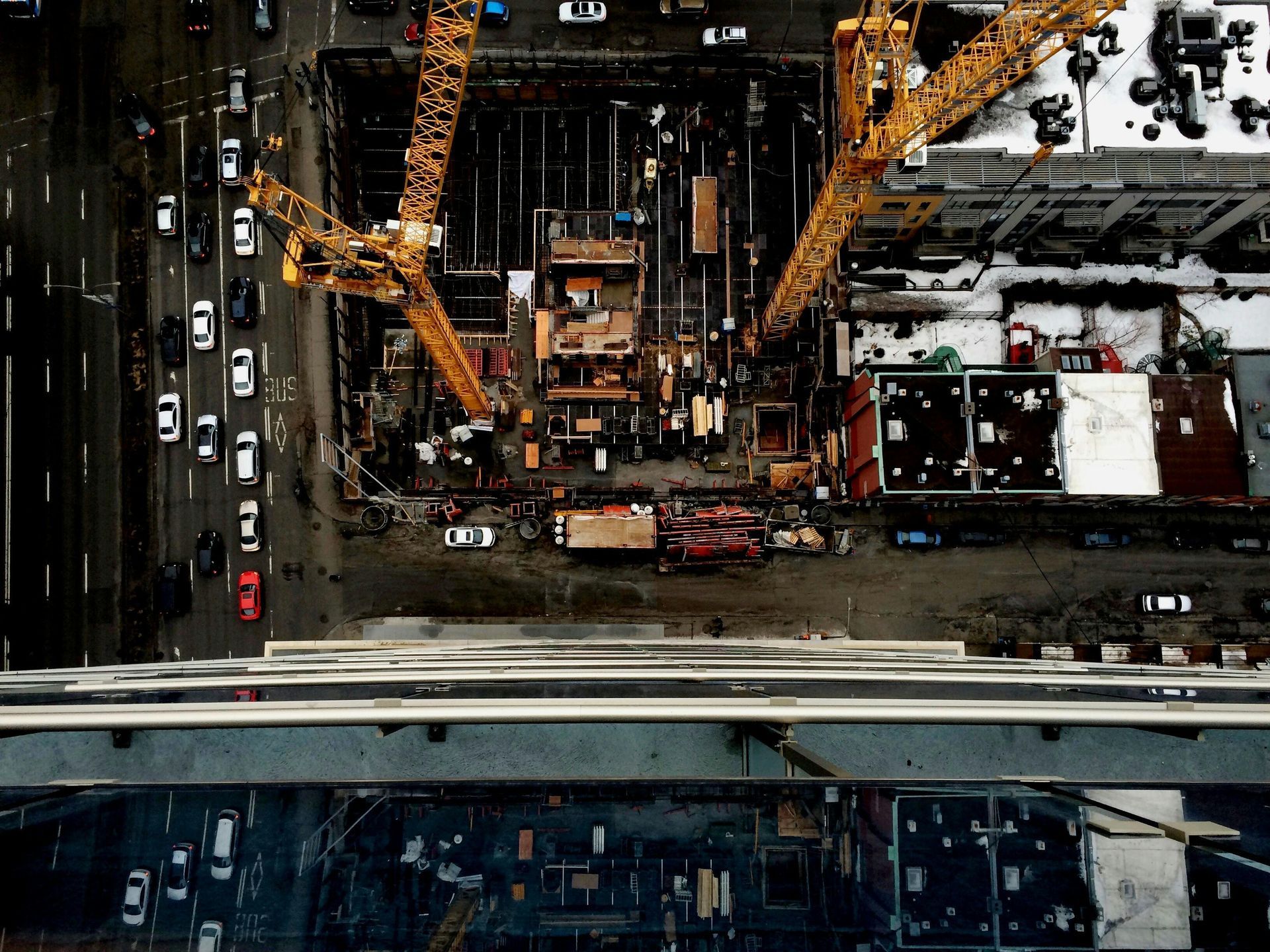 Overhead view of a construction site with cranes, buildings, and traffic on adjacent roads.