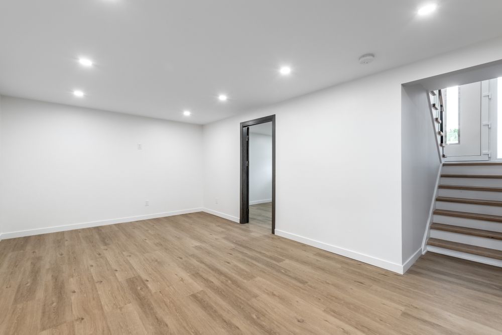 Empty room with hardwood floors, white walls, recessed lights, and a staircase.