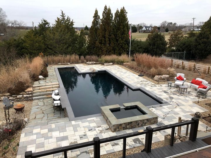 A dark blue rectangular pool and hot tub surrounded by stone patio, landscaping, and a distant lawn.