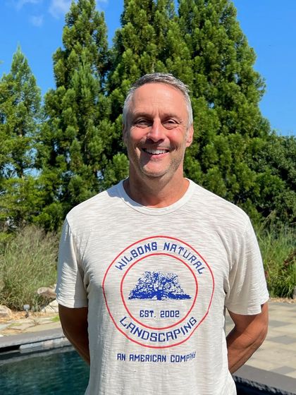 Man in white t-shirt, stands smiling near pool, tree background. T-shirt reads