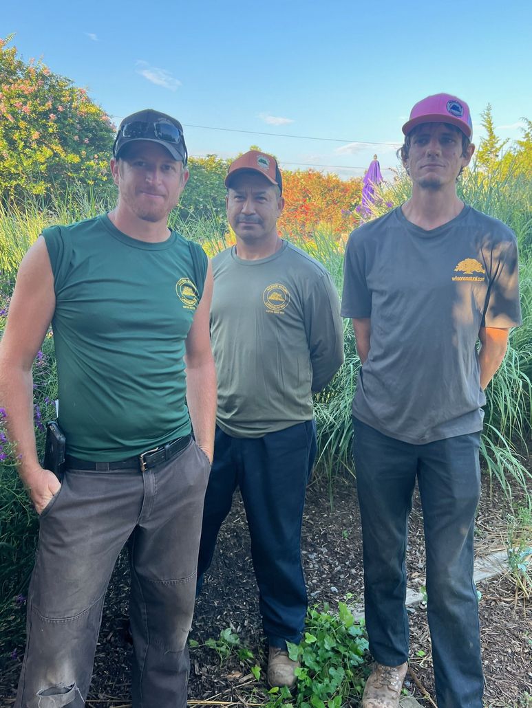 Three people in work clothes stand in a garden. The person on the left wears a green shirt and work pants. The middle person wears a shirt and dark pants. The person on the right wears a grey t-shirt and jeans.