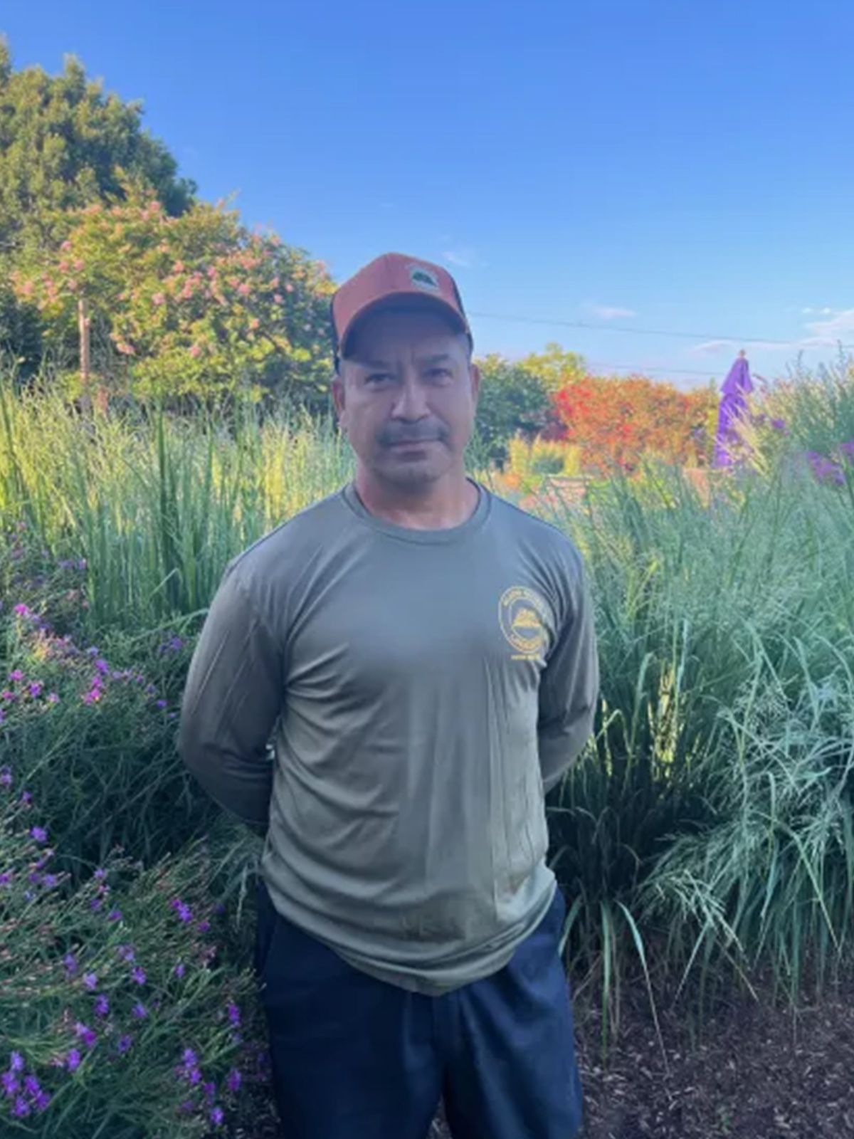 Man in a baseball cap and green shirt stands in a garden with tall plants and flowers, hands behind his back.