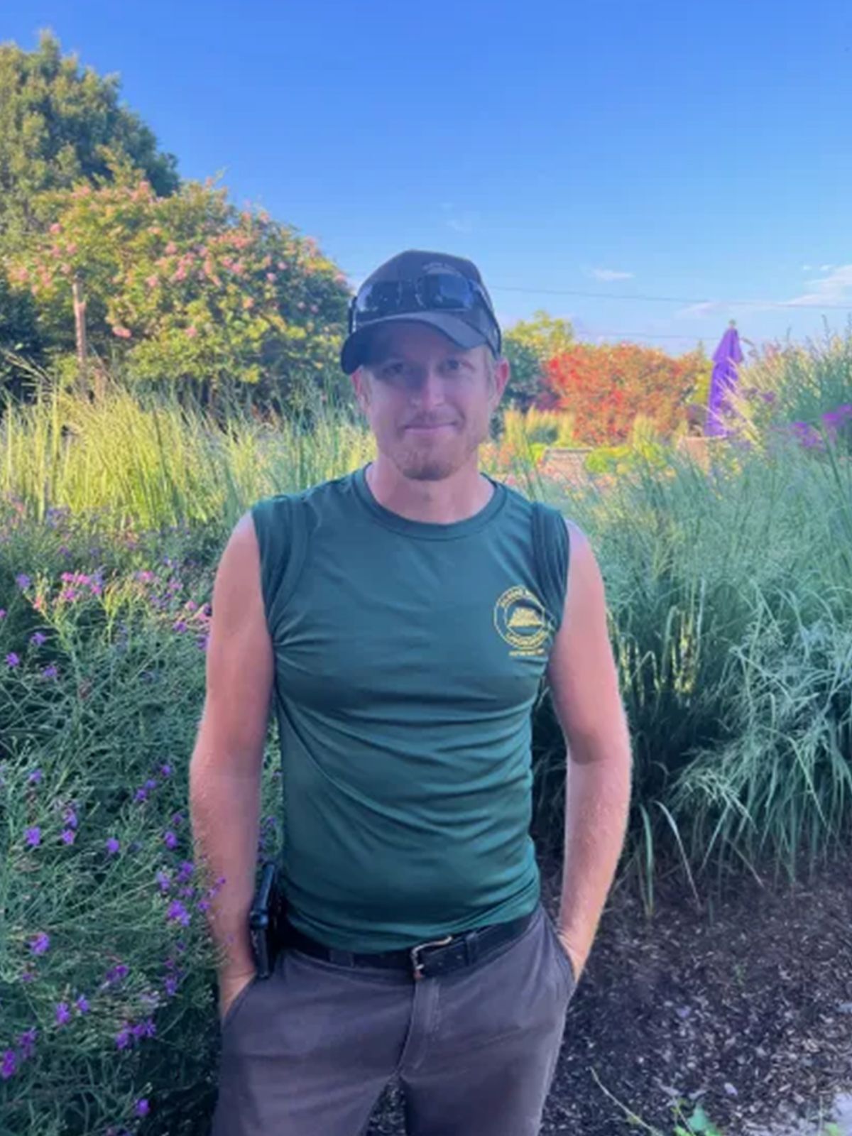 Man in green shirt, hands in pockets, stands in a garden with purple flowers and greenery; daytime.