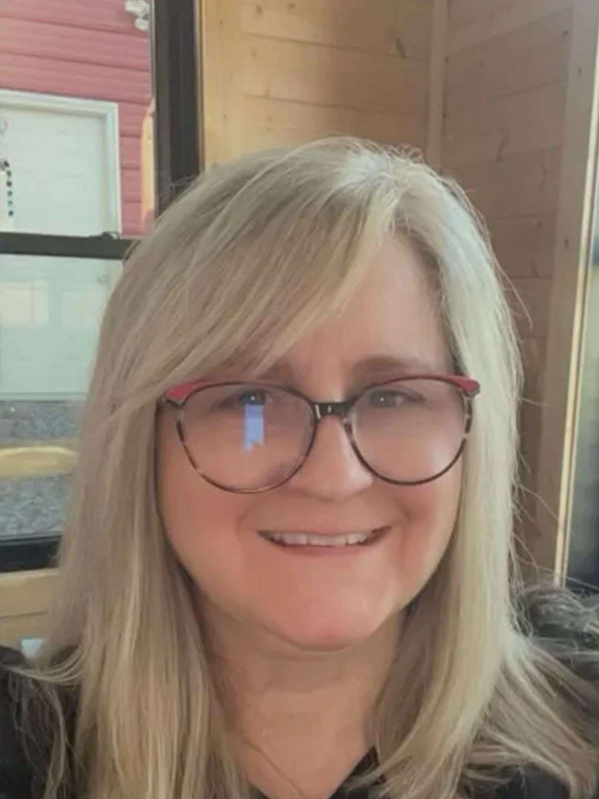 Woman with blonde hair and glasses smiles. Interior setting, wooden paneling.