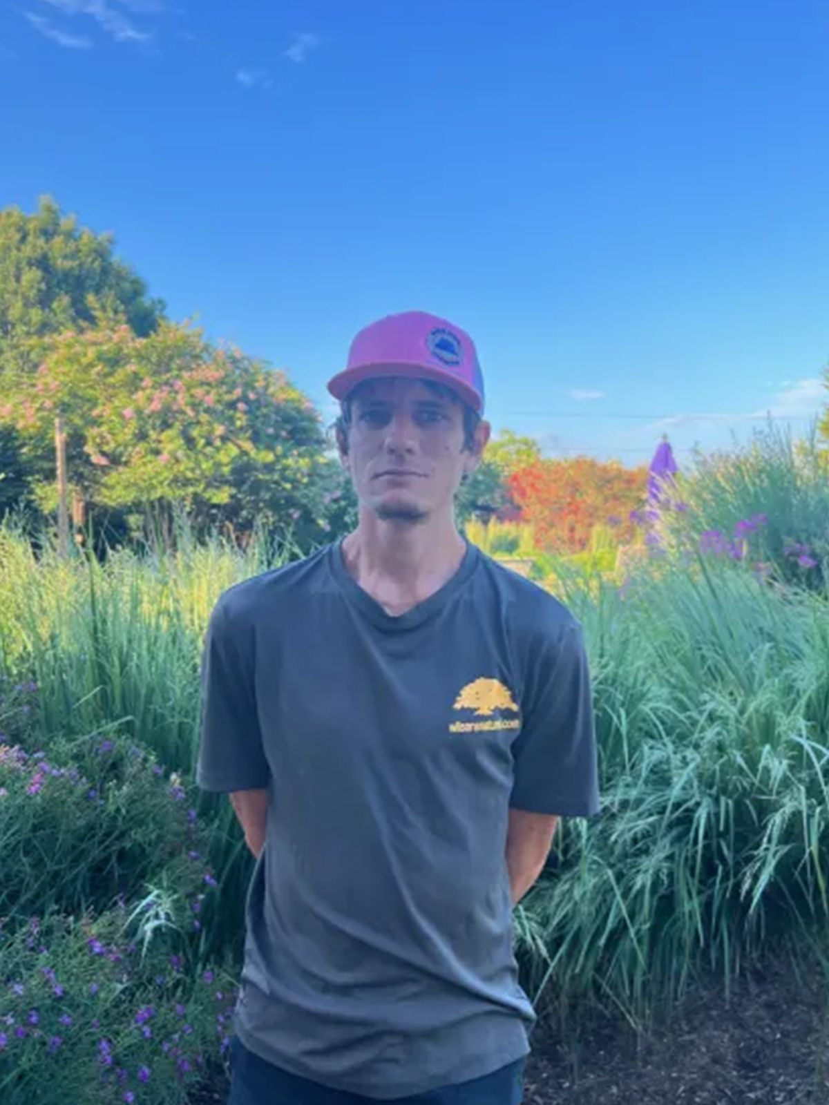 Man in gray t-shirt and pink cap stands outdoors in a garden, hands clasped behind back.
