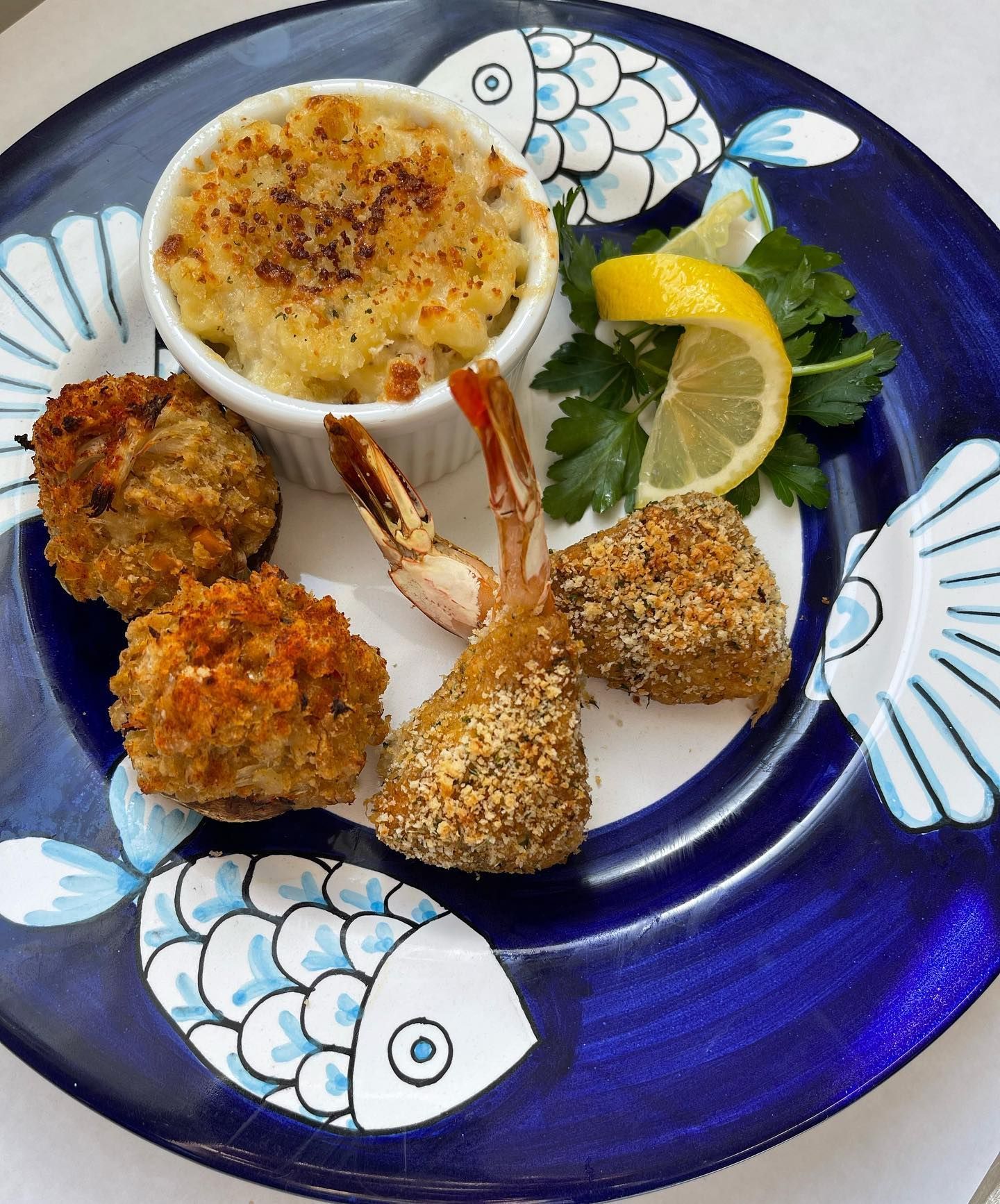 Seafood appetizer plate: mac and cheese, fried shrimp and scallops, lemon slice, and parsley on a blue fish-decorated plate.