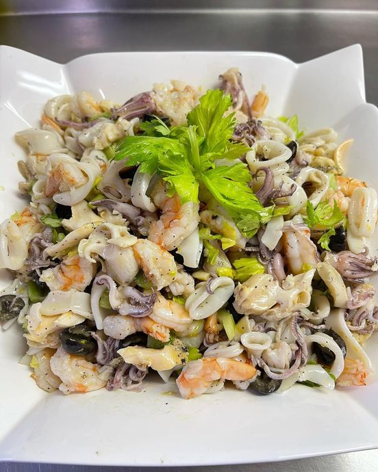 Seafood salad in a white bowl, featuring shrimp, squid, and olives, garnished with celery.