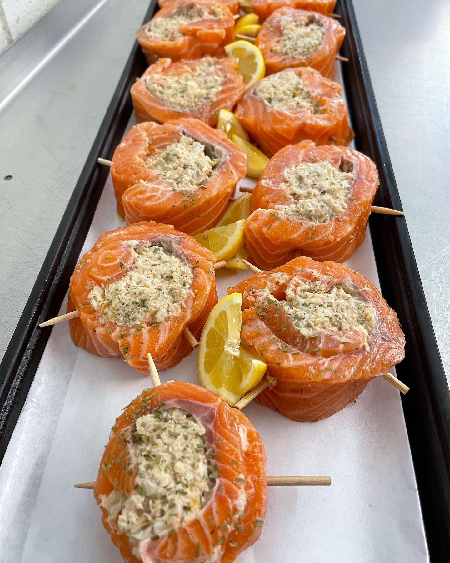 Salmon rolls stuffed with filling, secured with skewers, garnished with lemon wedges, in a tray.
