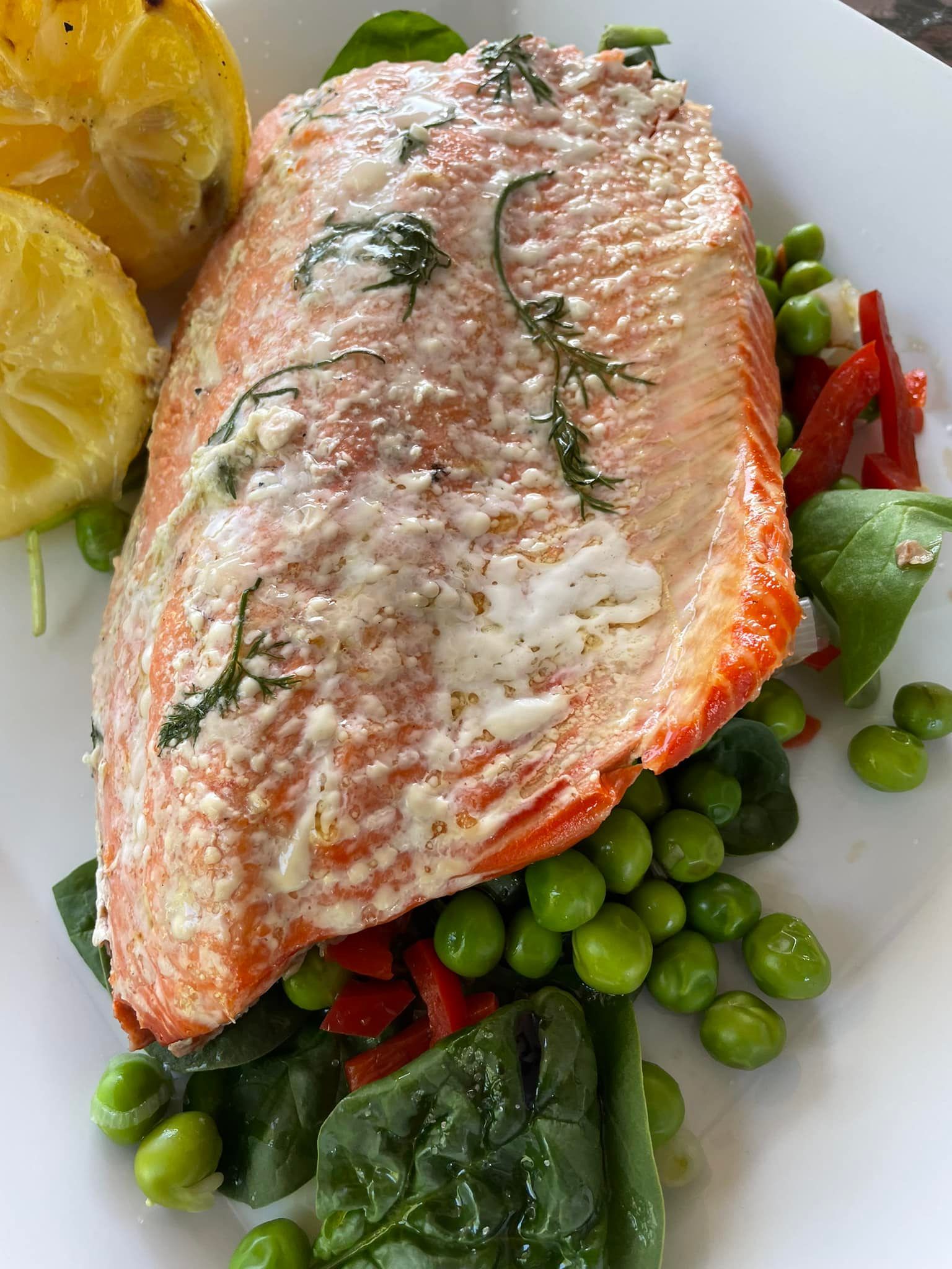 Salmon fillet with creamy sauce, peas, spinach, red peppers, and lemon on a white plate.