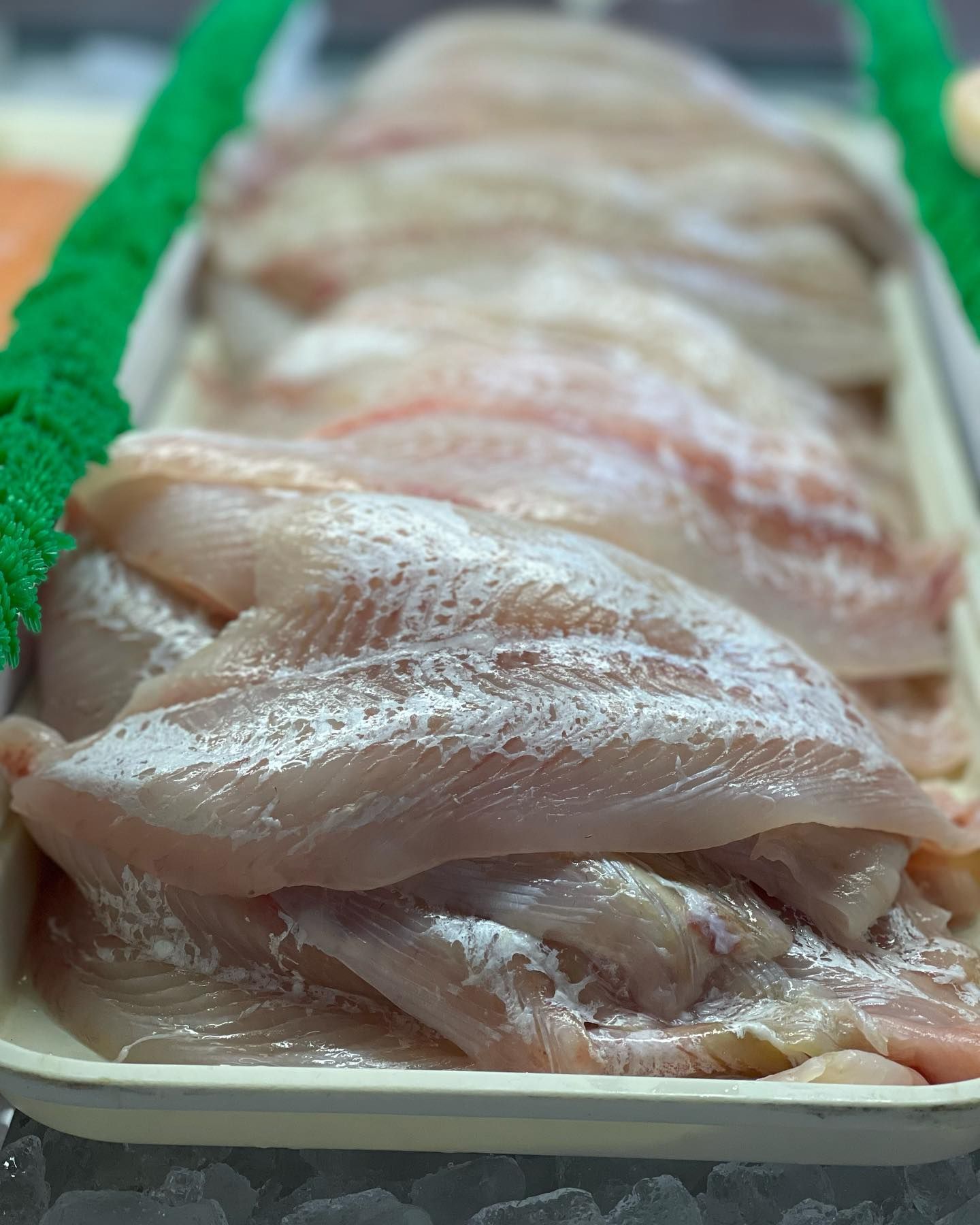 Raw, white fish fillets in a white tray at a seafood counter, with a green decoration on the side.