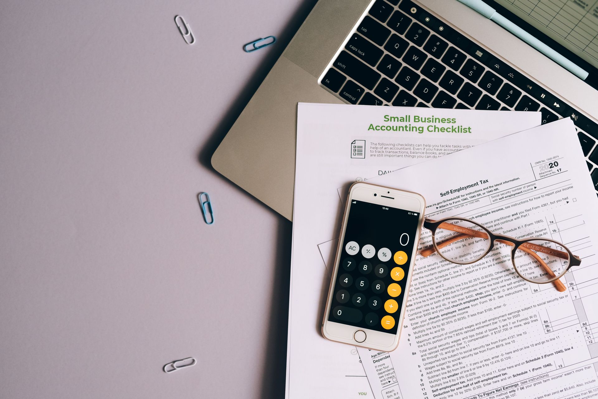 Photo of laptop, documents, calculator, glasses, and paperclips on a grey table