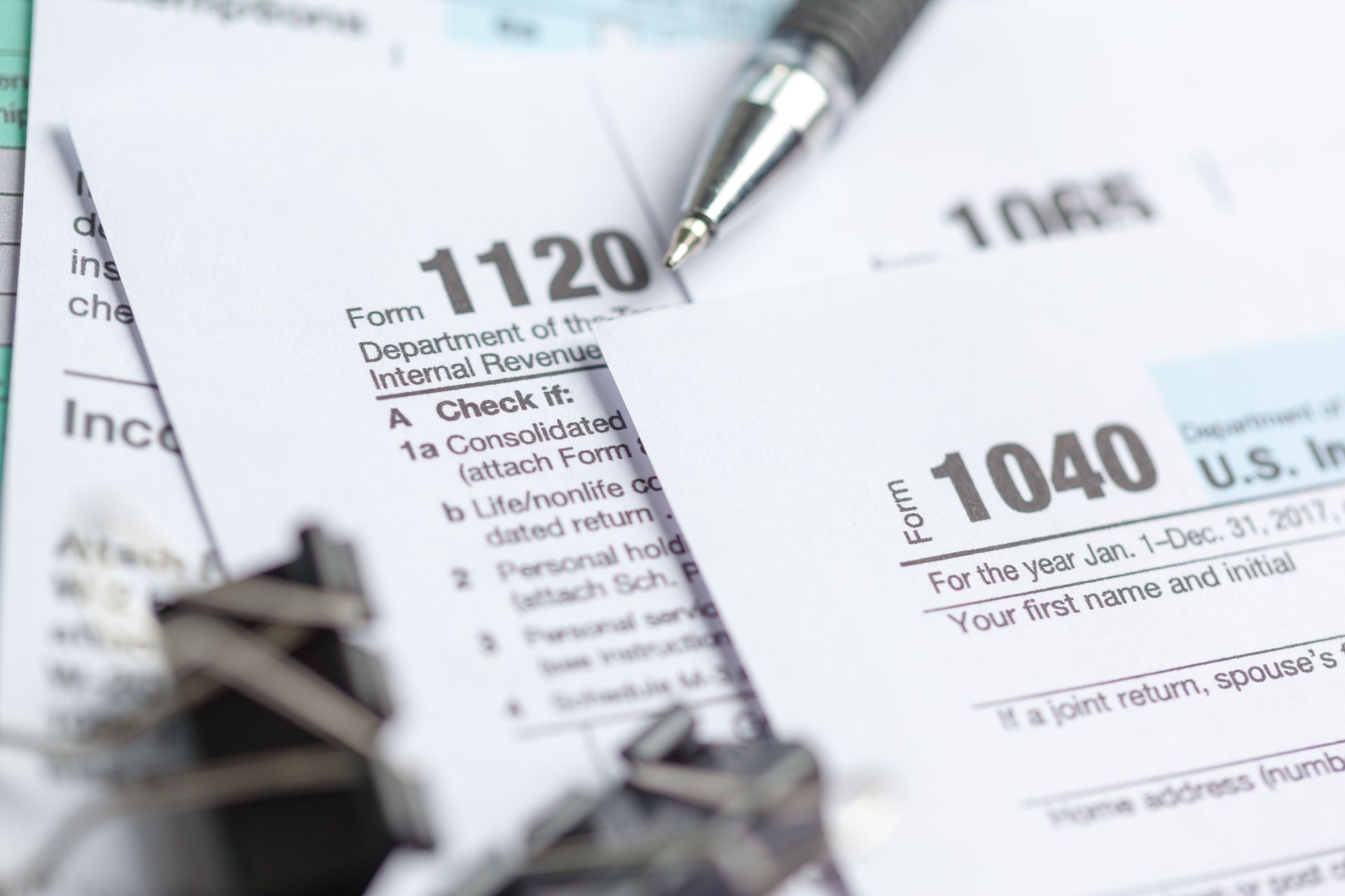 A close-up of U.S. tax forms 1120, 1040, and 1065 arranged on a surface with a pen and binder clips.