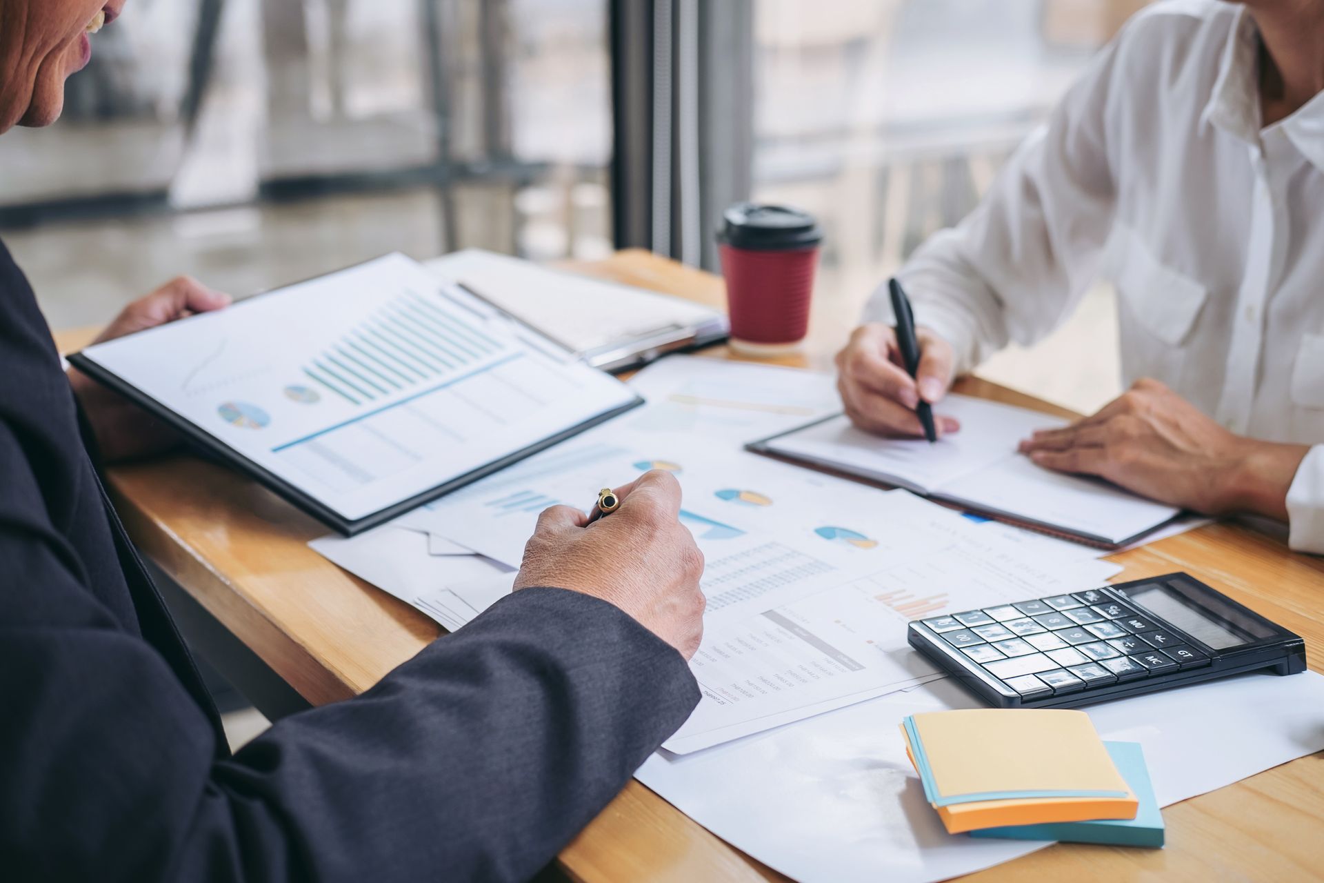 Two professionals in business attire review financial charts and documents at a desk with a calculator and coffee.