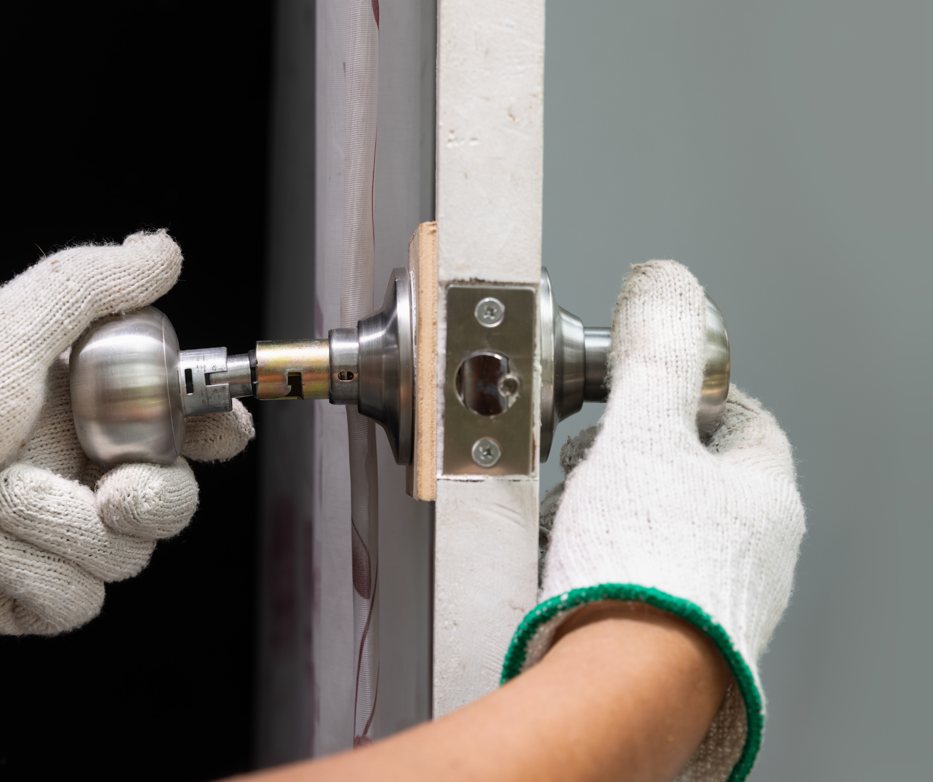Hands in work gloves installing a silver doorknob on a white door frame.