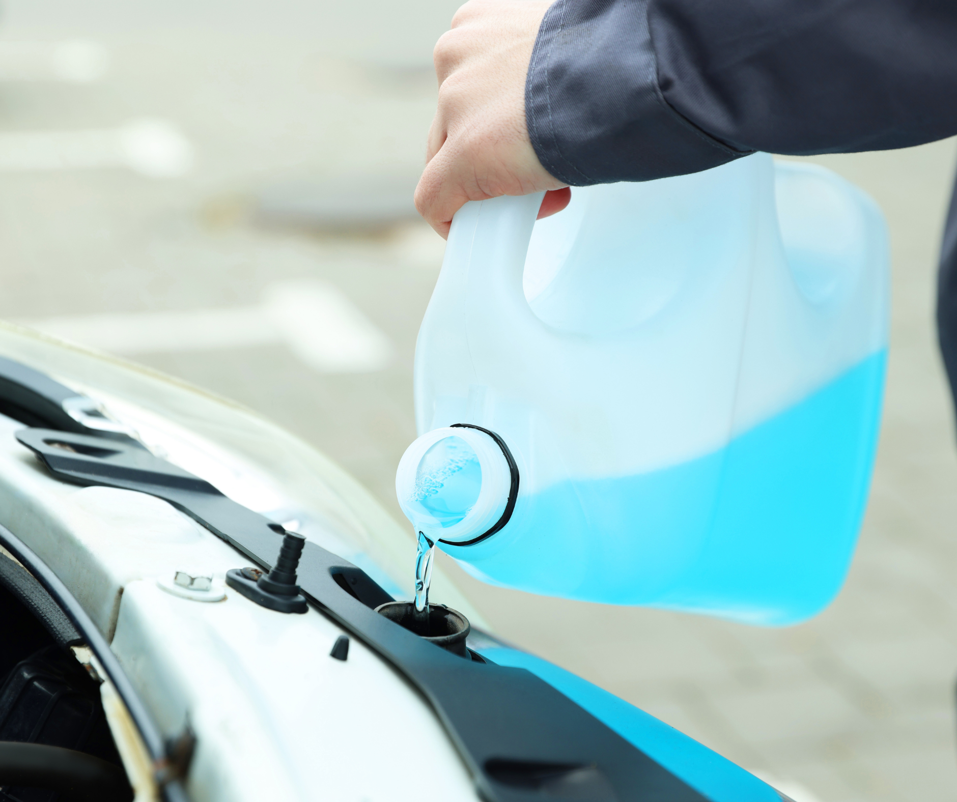 Person filling a car's windshield washer fluid reservoir with blue liquid from a white container.