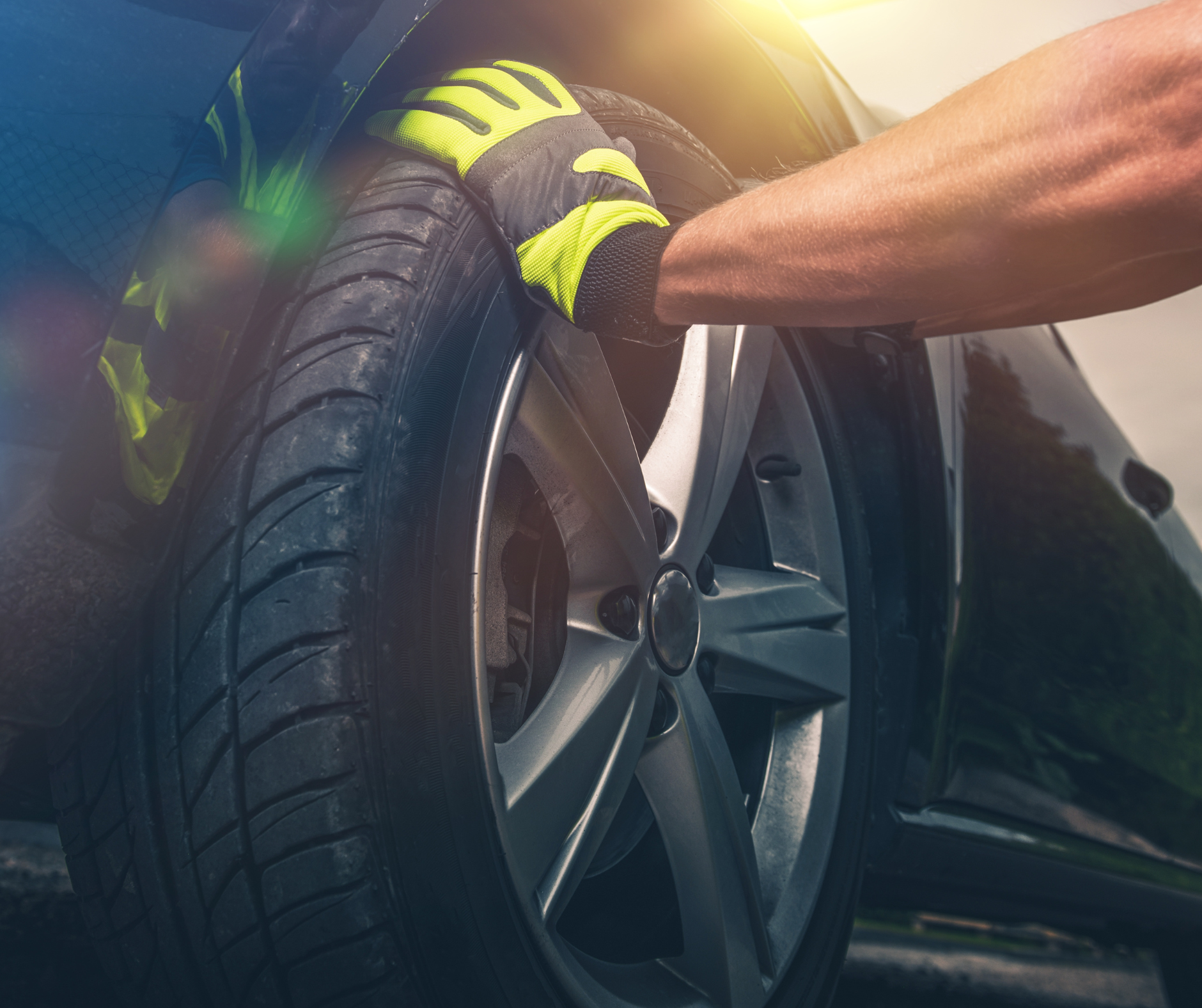 Person wearing yellow gloves checking a car tire on a sunny day.