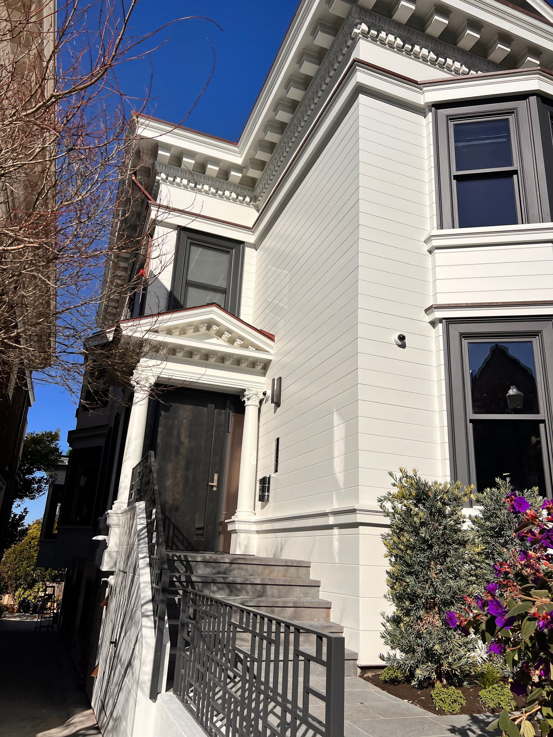 White Victorian-style house with ornate trim, black iron railings, and steps beside purple flowers under a blue sky