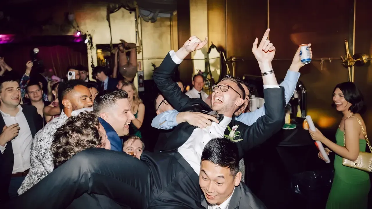 People celebrating, carrying a man aloft in a dimly lit room; arms raised, expressions of joy.